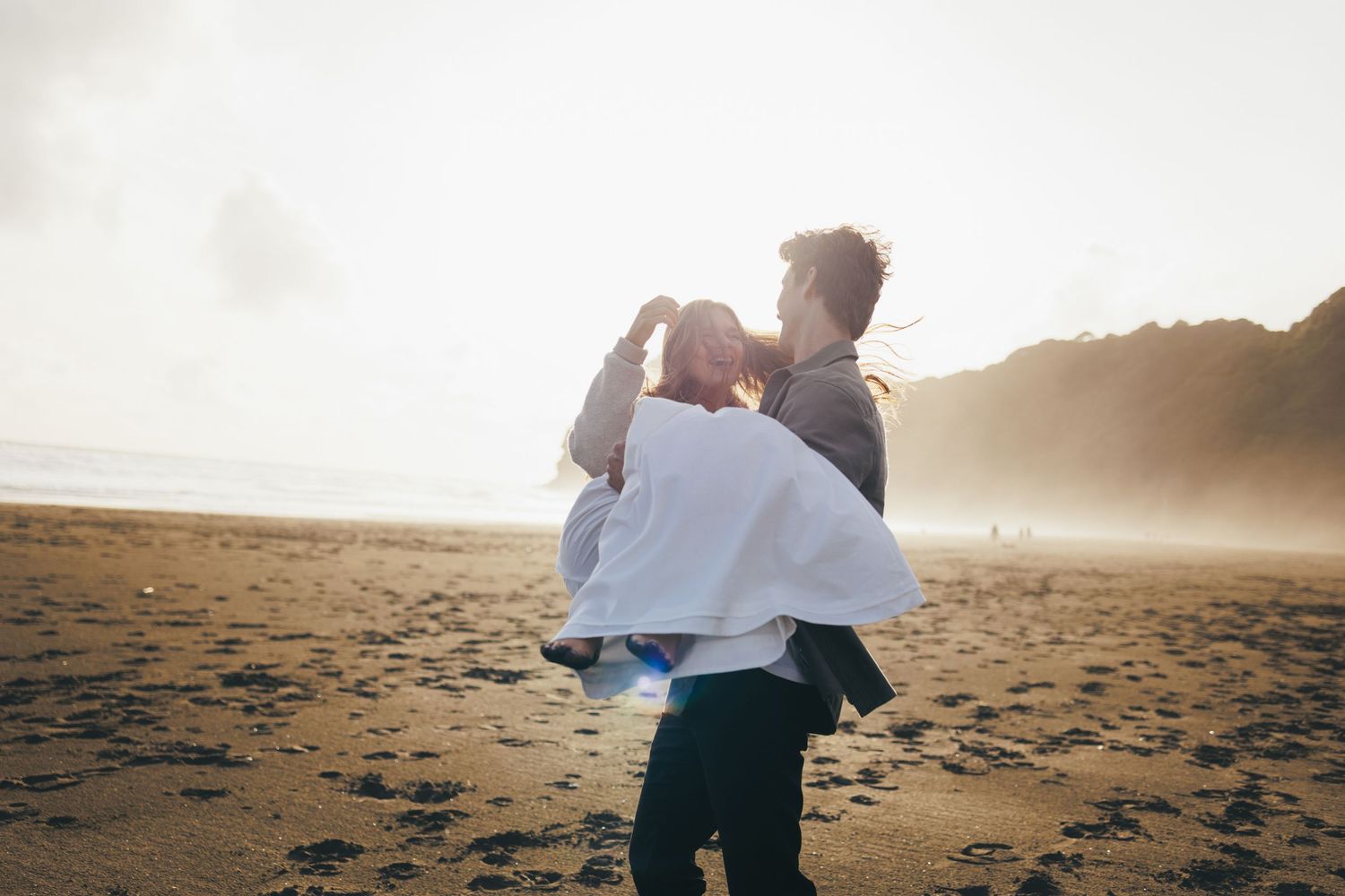 Man lifting and spinning woman on a sunny beach with misty cliffs in background.