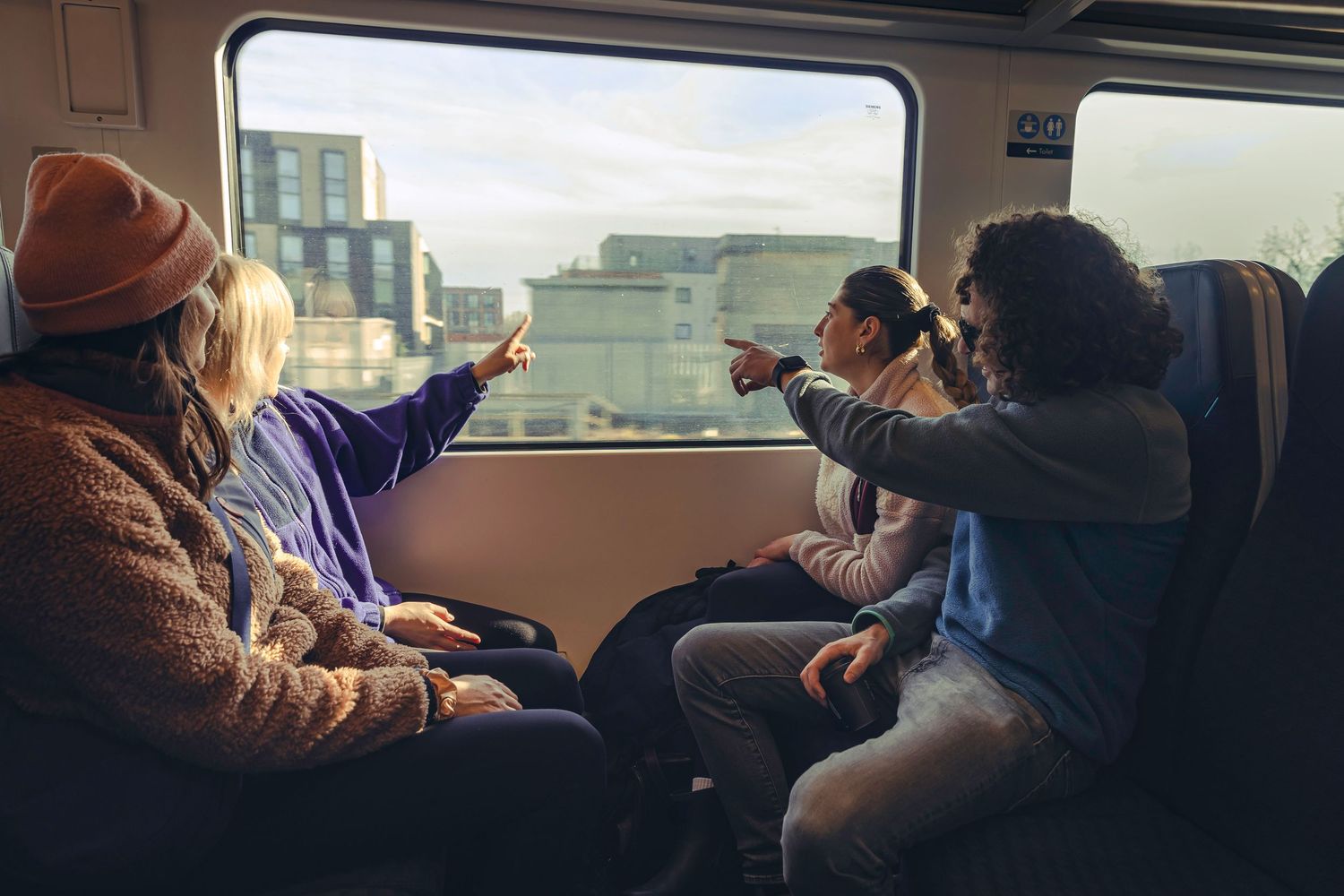 Group of friends pointing out the window at city views while riding on a sunny train journey.