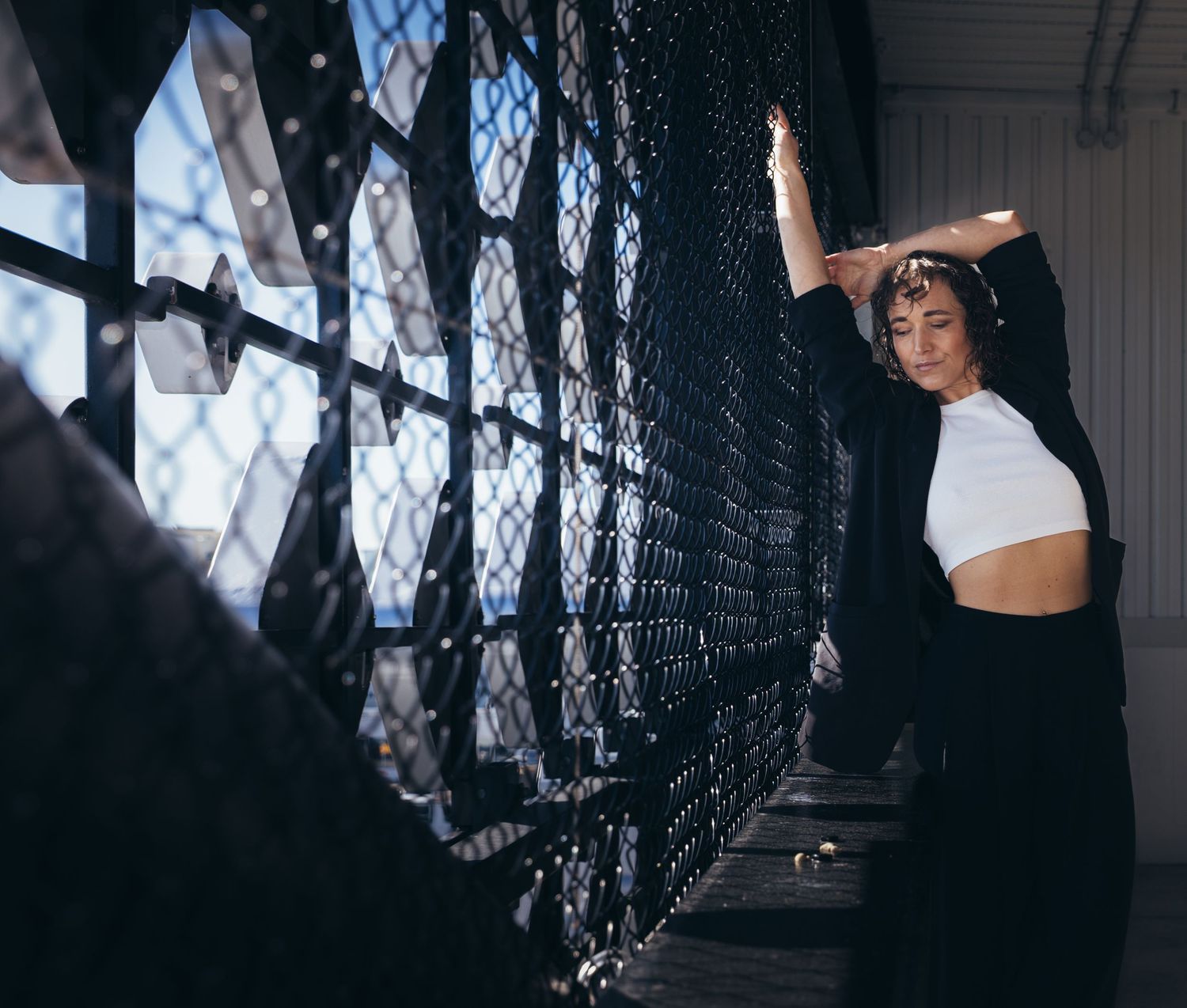 Woman in white crop top and black jacket leaning against decorative metal fence in dramatic lighting.