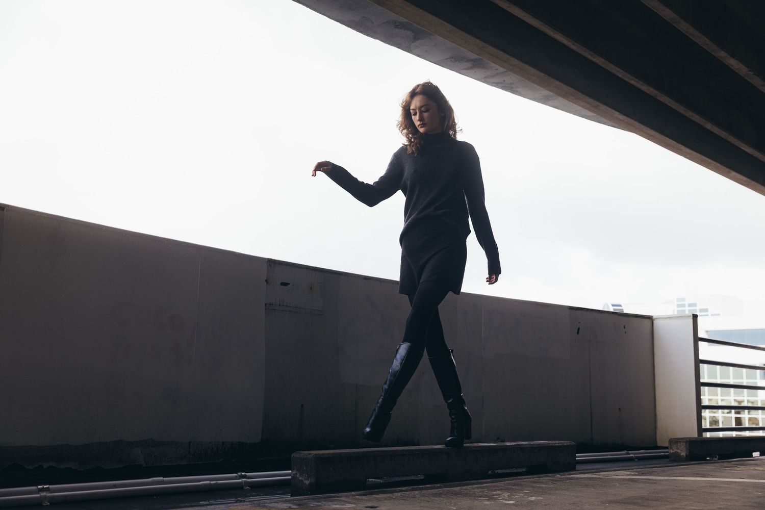 Woman in black turtleneck and boots walking on ledge in urban concrete underpass environment.