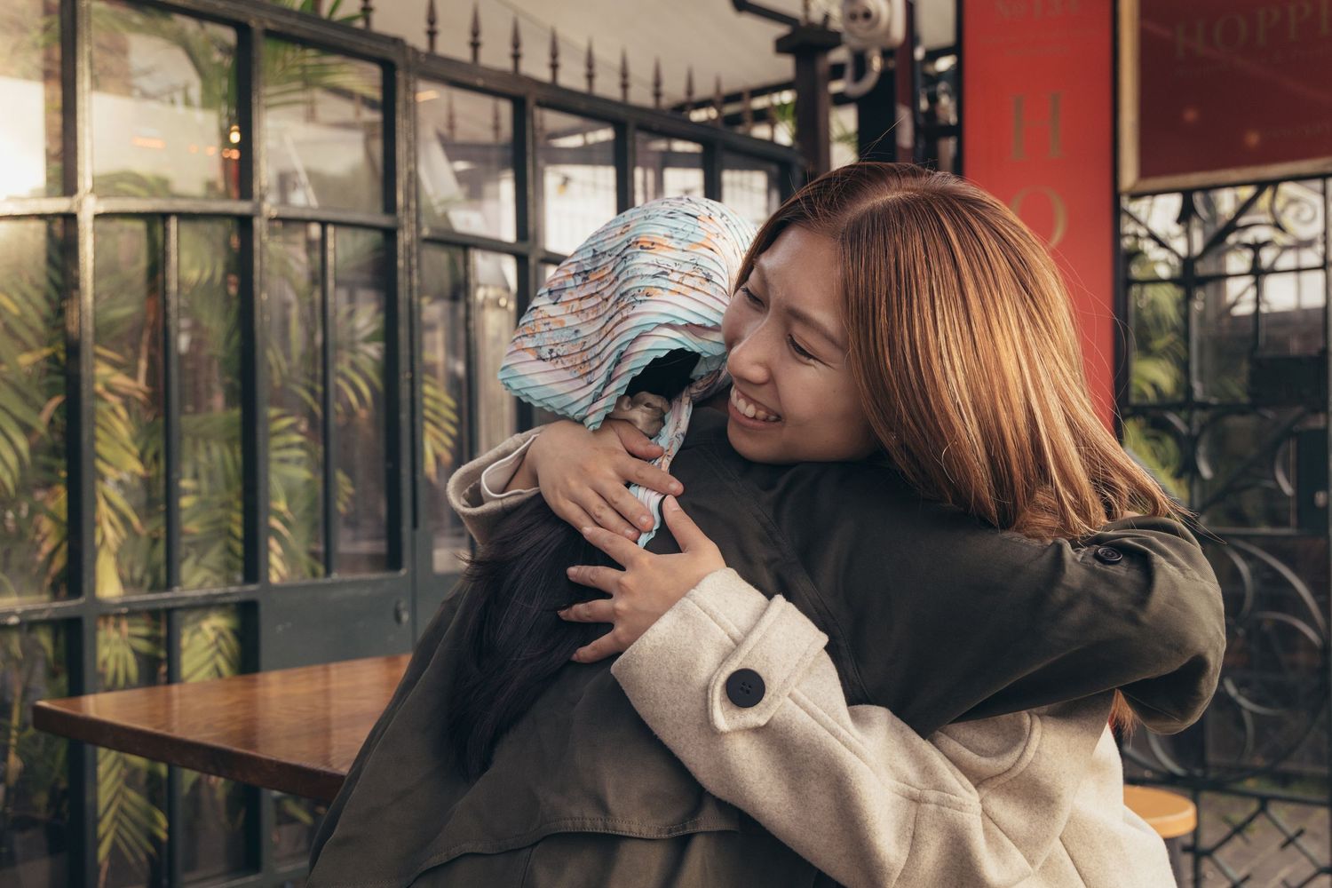 Two women sharing a warm hug inside a cozy cafe, one wearing a colorful beanie hat.