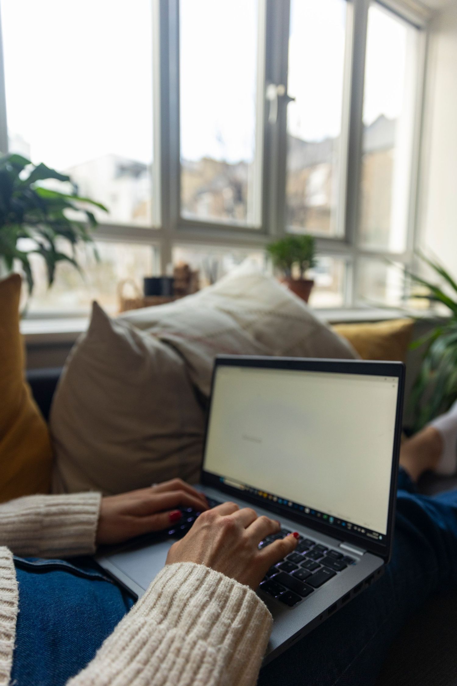 Person in cream knit sweater typing on laptop while relaxing on couch near bright window.
