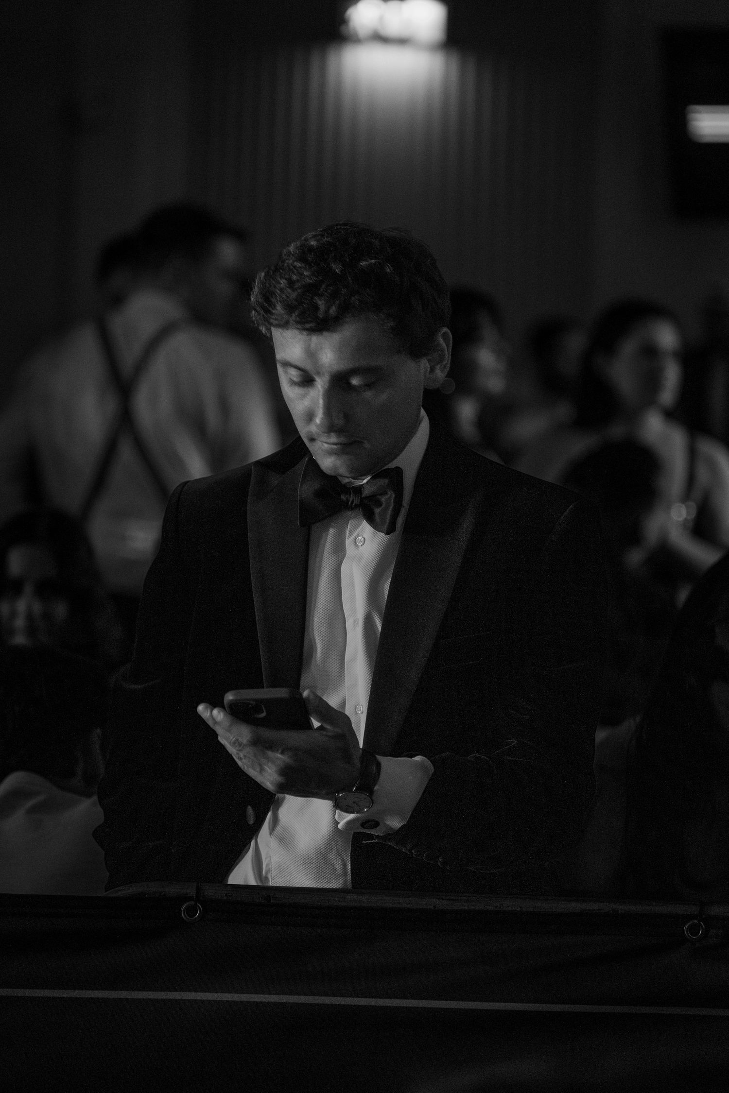 A young man in a tuxedo and bow tie looks at his phone at a formal evening event.
