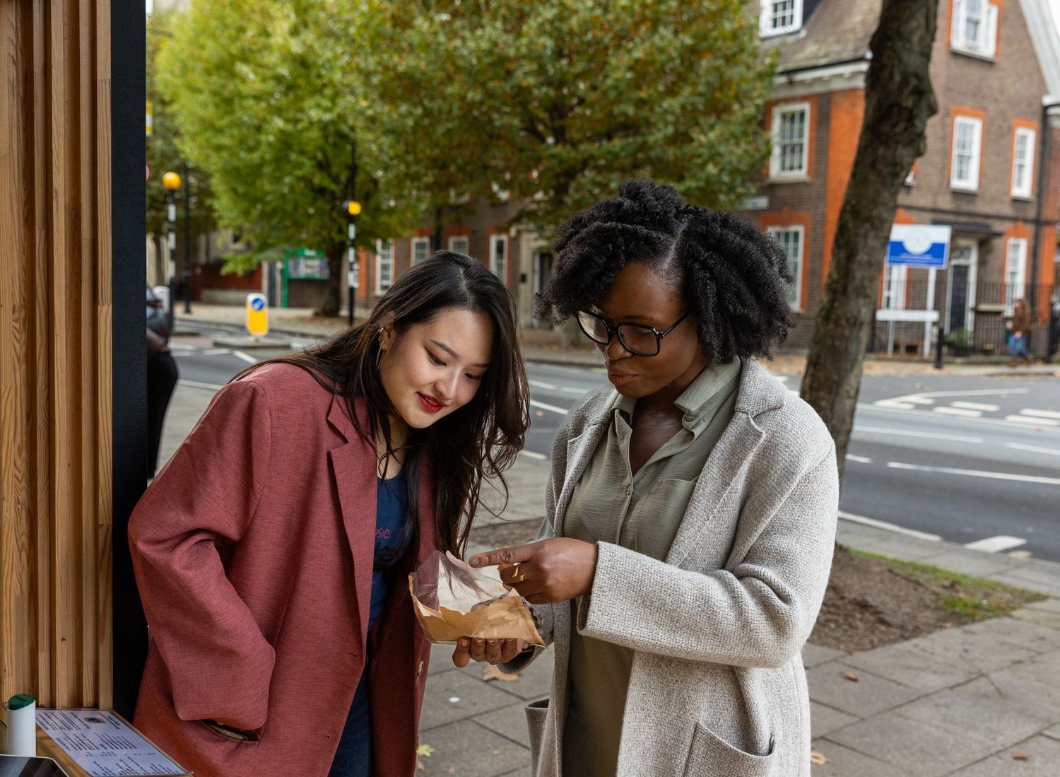 Two young women examining food together on a London street with trees and red brick buildings behind.