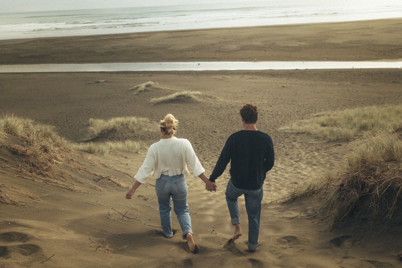 Couple holding hands walking barefoot down sandy beach dunes toward the ocean shoreline.