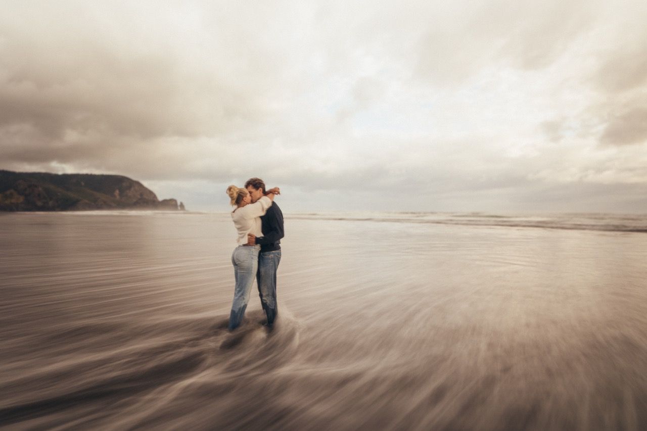 Couple embracing and kissing on a windy beach with dramatic cloudy skies and coastal cliffs in the background.