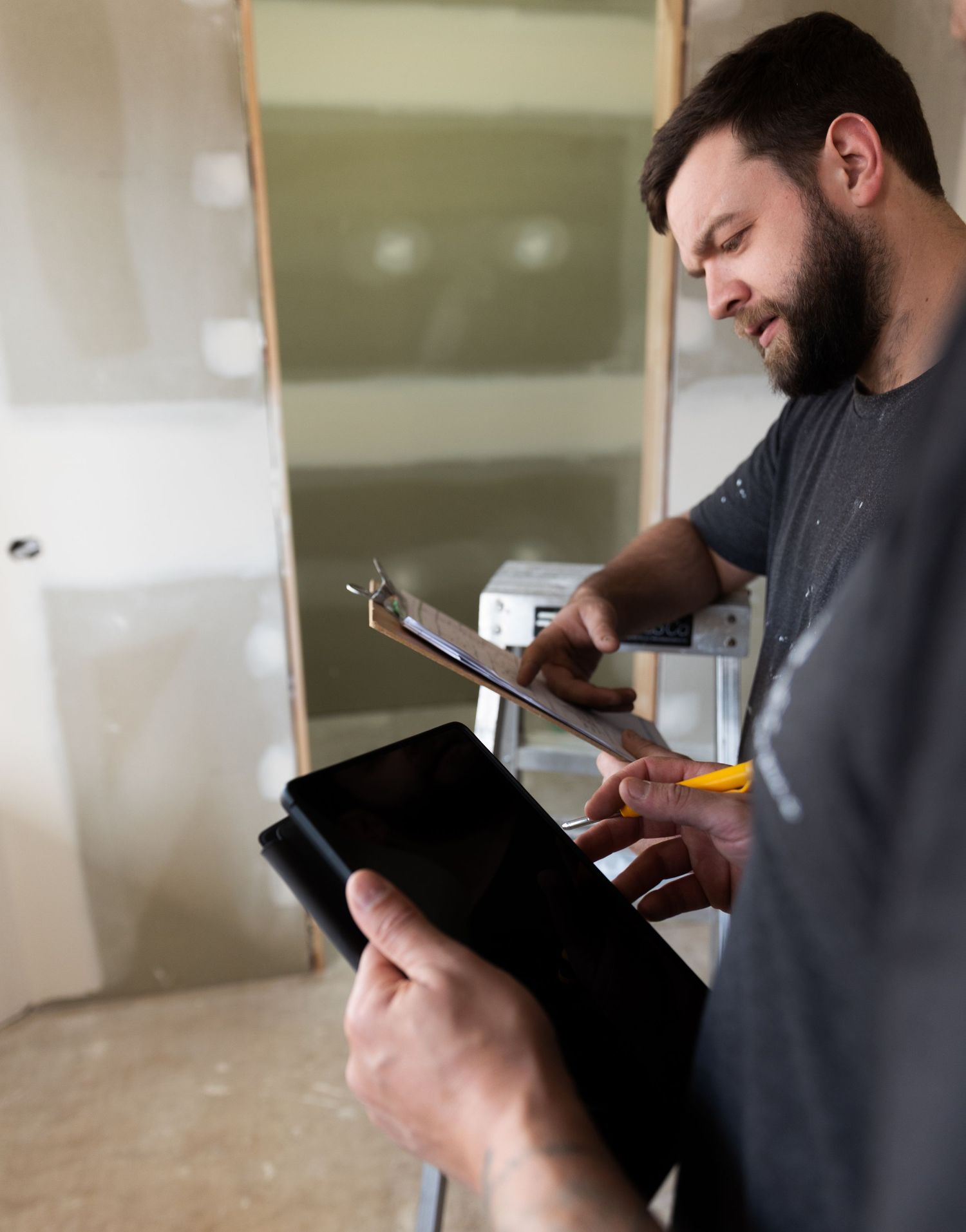 Bearded builder smiling while reviewing clipboard blueprints and holding a digital tablet.