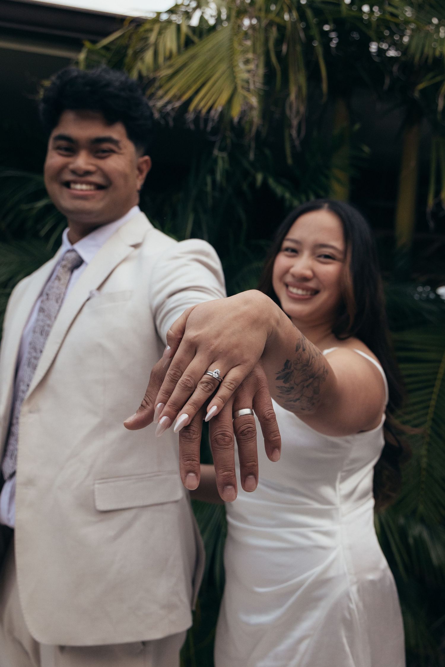 Newlywed couple in white wedding attire smiling while showing off their wedding rings outdoors with tropical greenery.