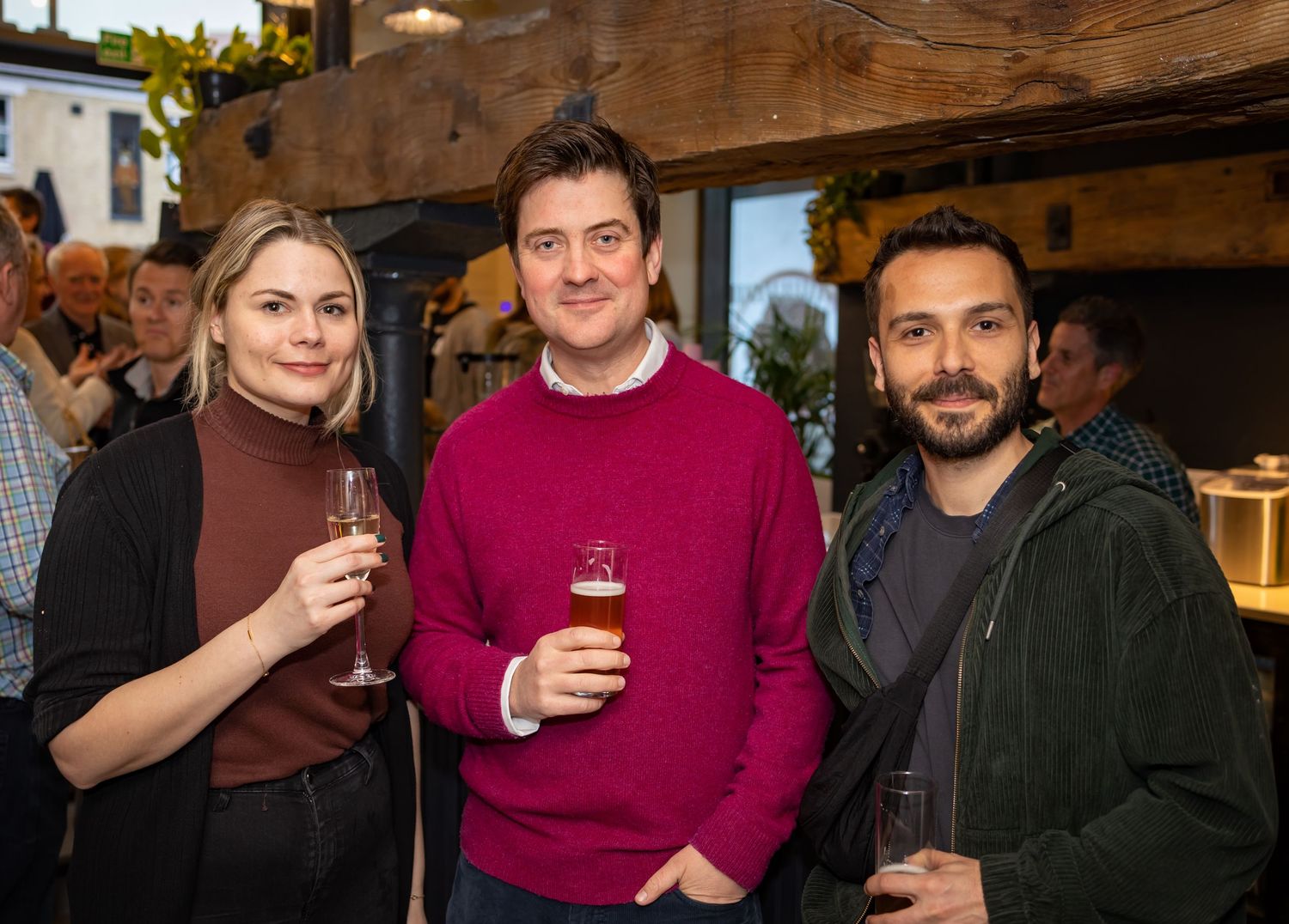 Two women and a bearded man smile together holding drinks at an indoor networking event.