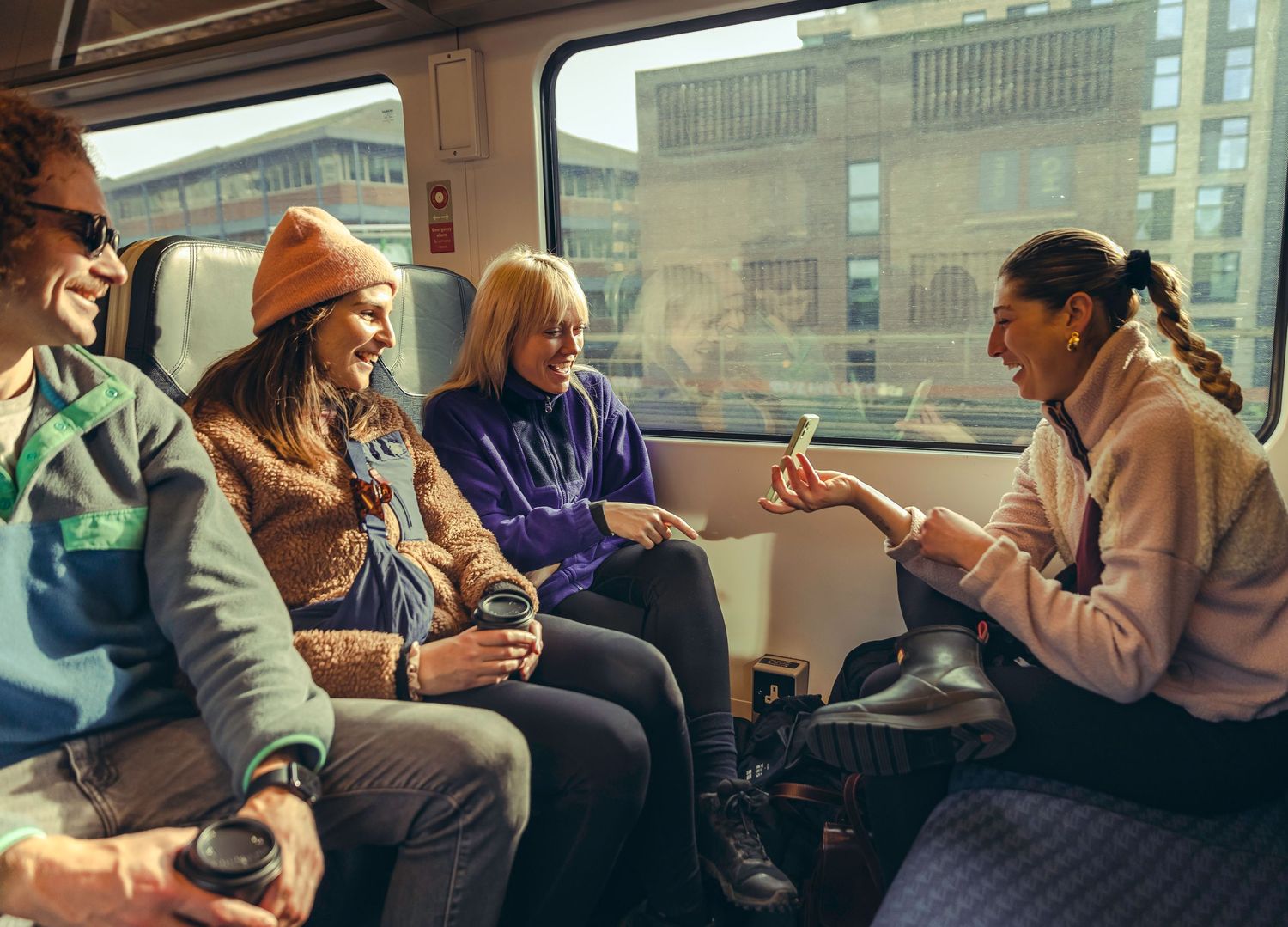 Four friends laughing and talking together while seated on a train passing urban buildings.