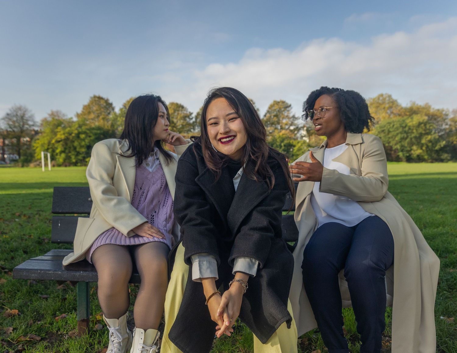 Three women sitting on a park bench laughing and enjoying time together on a sunny autumn day.