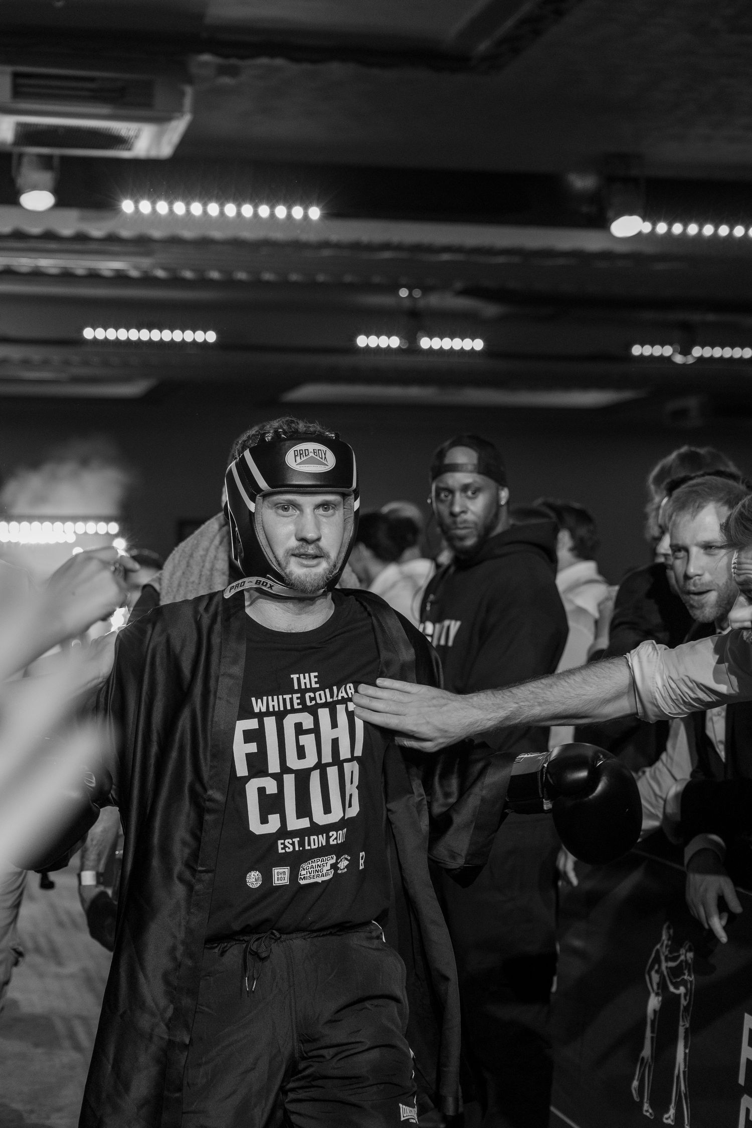 A man in a White Collar Fight Club t-shirt walks through a crowd at a boxing event.