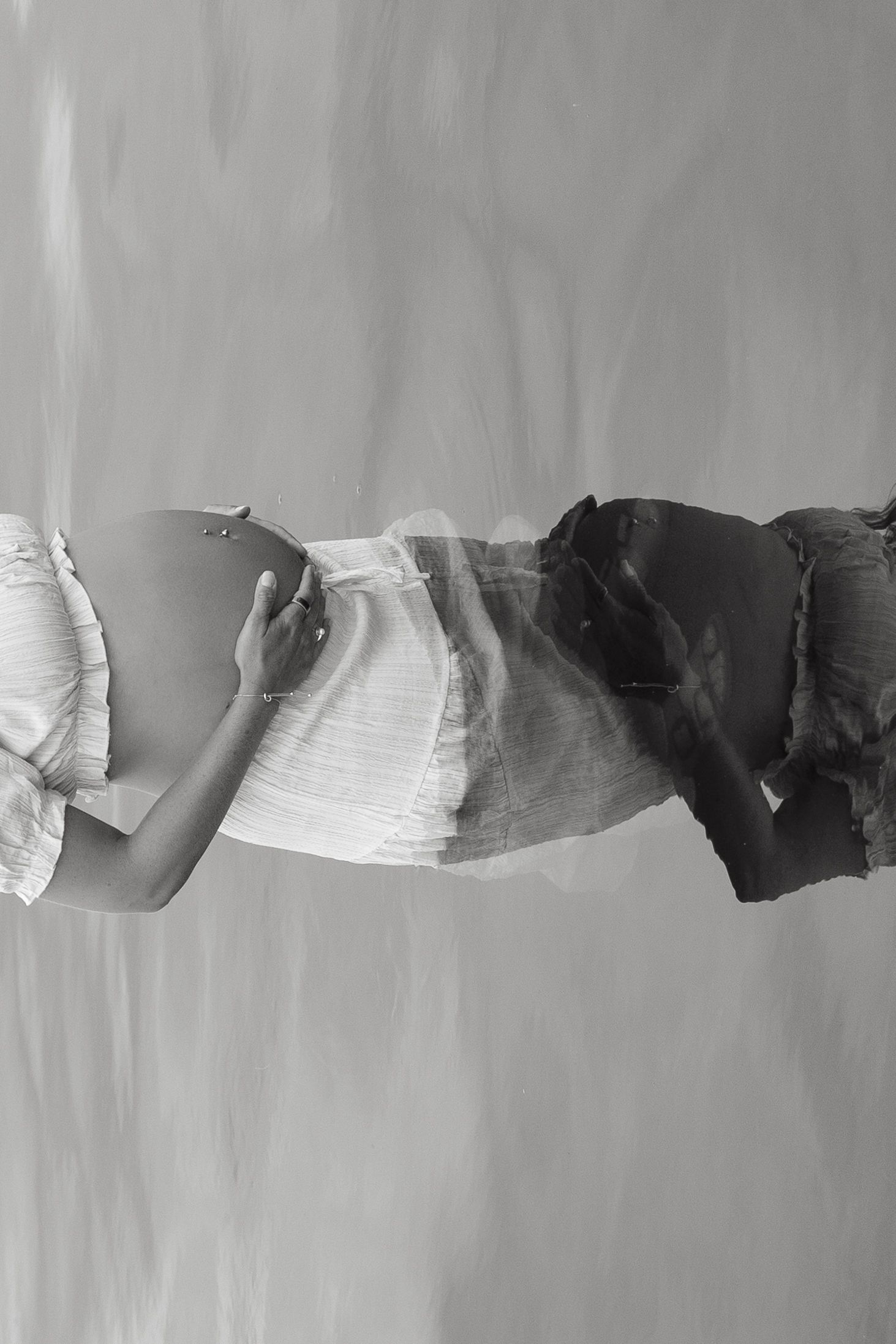 Black and white double exposure photo of a pregnant woman cradling her baby bump in a white dress.