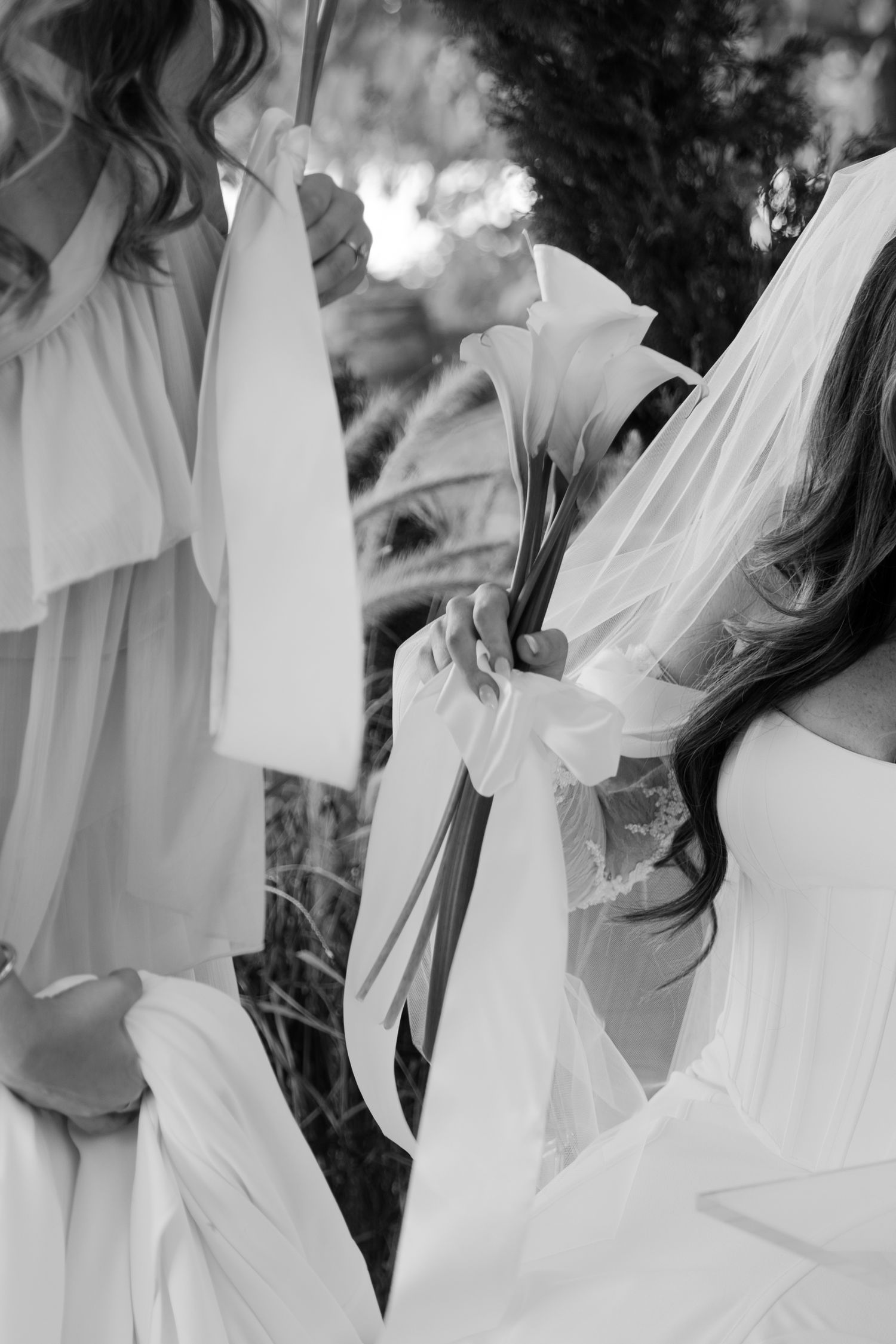Close-up black and white photo of bride holding floral bouquet with ribbon in outdoor wedding setting.