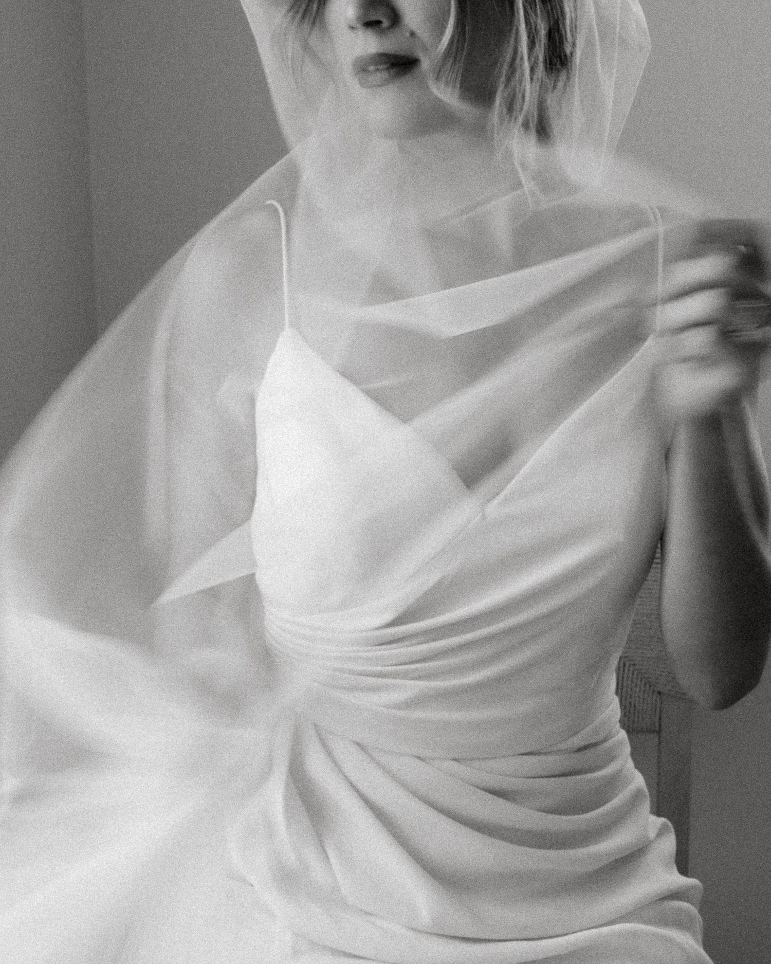 Black and white photo of bride in slip gown holding sheer veil across chest in artistic composition.