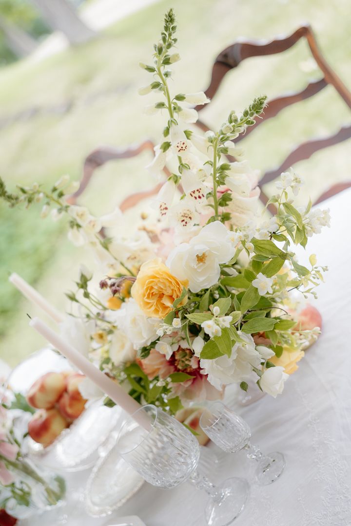 Elegant wedding centerpiece with white and yellow roses, foxglove, and greenery in crystal vase on white tablecloth.
