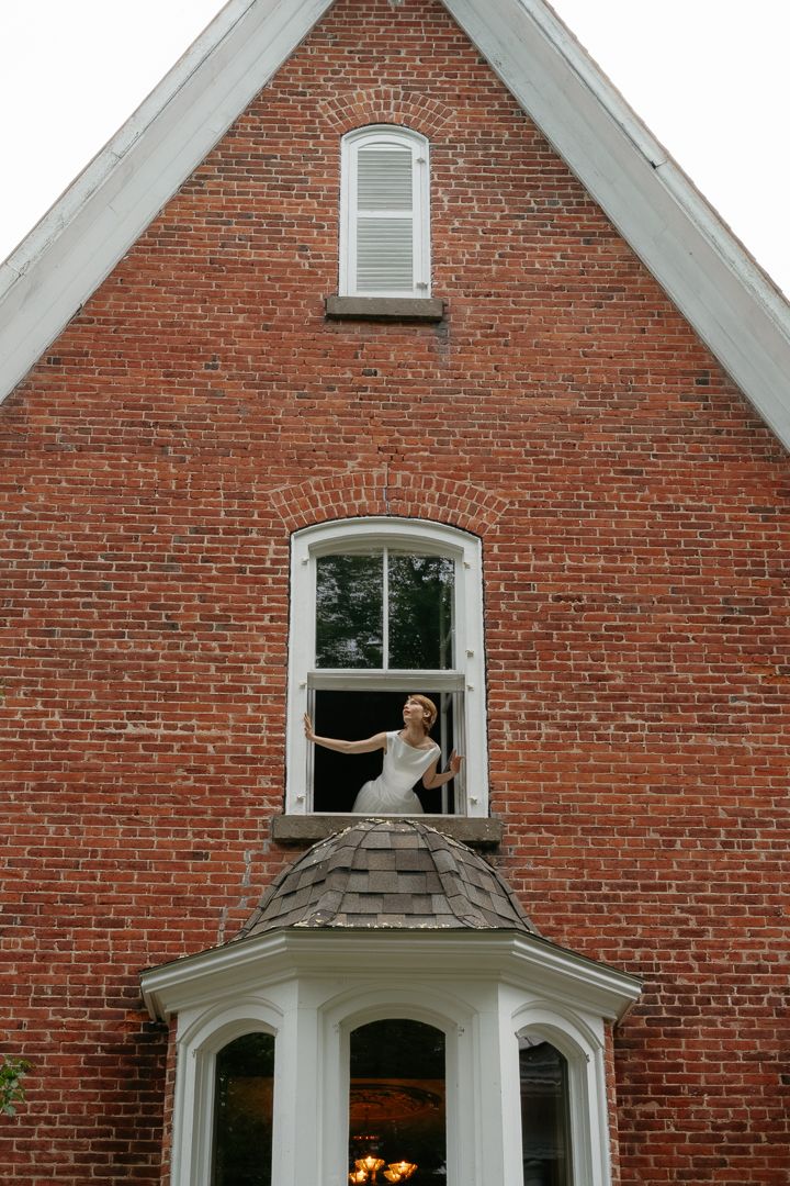 Bride in white gown leaning out of an arched window on a red brick building with a gabled roof.