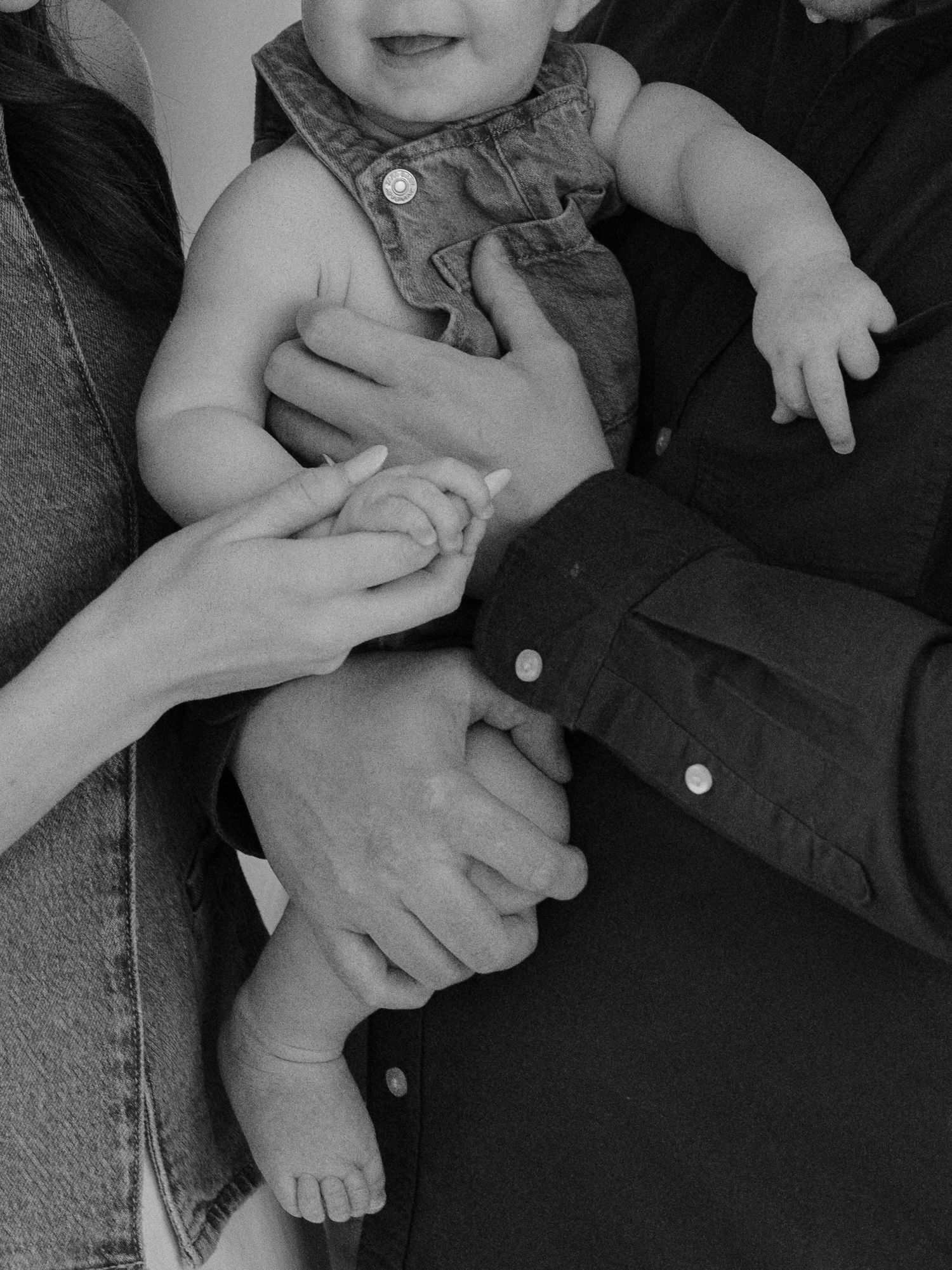 Black and white photo of parents holding their smiling baby in denim overalls, hands intertwined in a tender family moment.