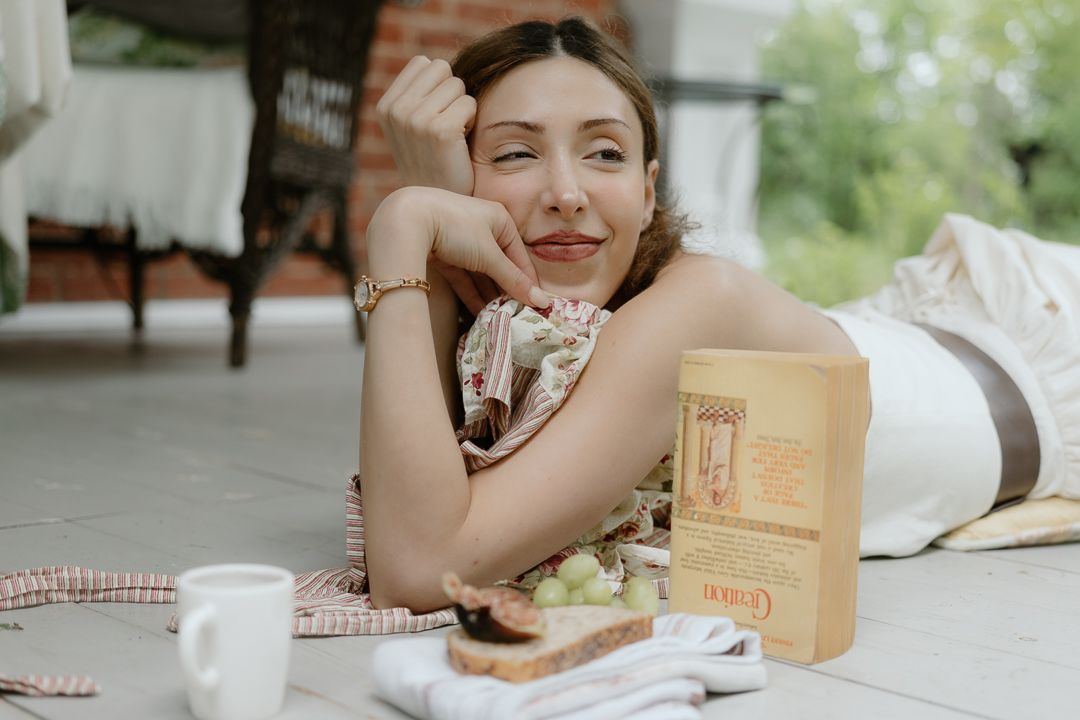 Smiling woman lying on a patio floor beside a book, grapes, toast, and a white coffee cup outdoors.