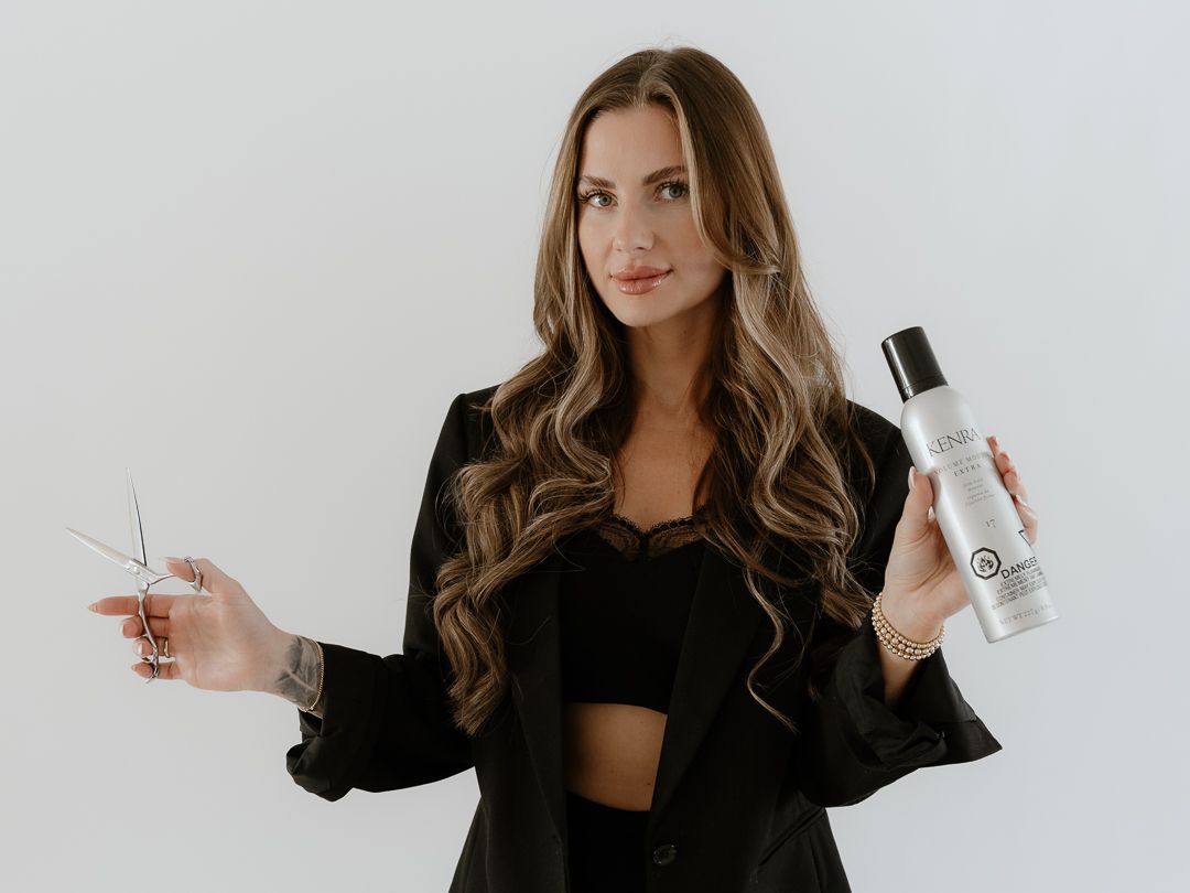 Woman in black blazer holding hair scissors and a white bottle of hair product against a light gray background.