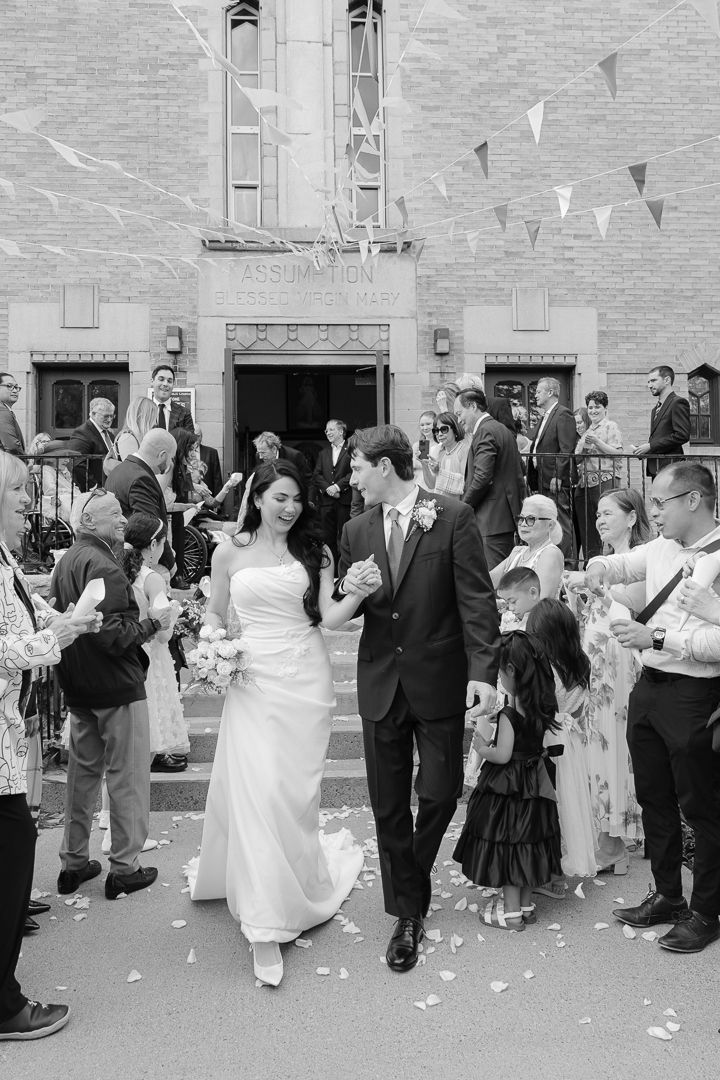 Black and white photo of a bride and groom exiting a church, surrounded by cheering guests throwing confetti.