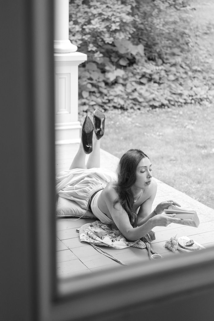 Black and white photo of a woman lying on a porch reading a book, with heels up and greenery in background.