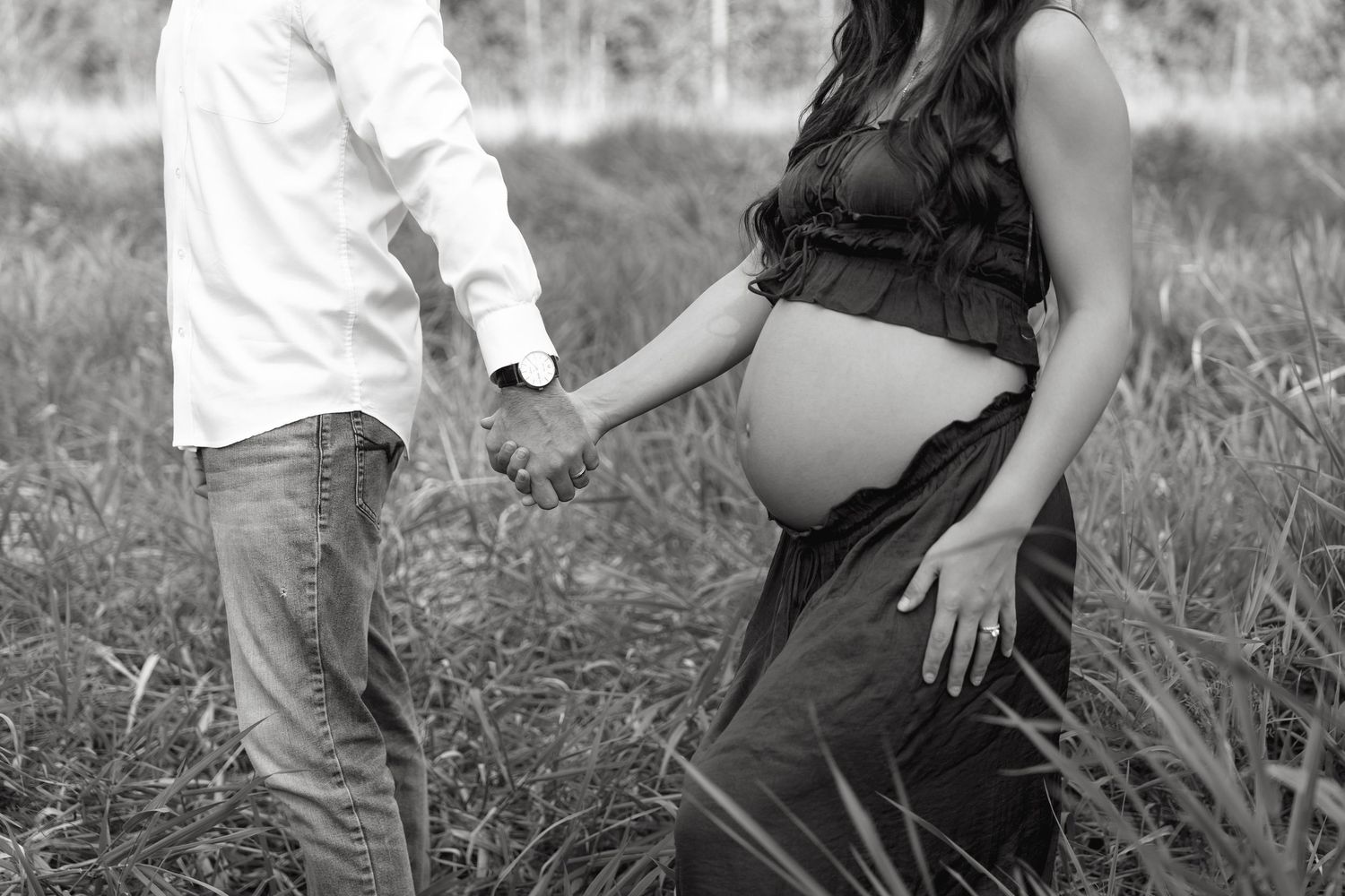 Black and white photo of a pregnant woman holding hands with her partner in a field, showcasing her baby bump.