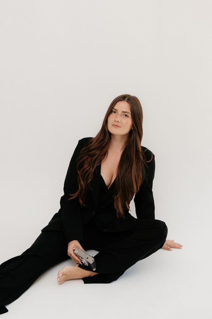 Woman in black suit sitting on white floor holding a camera, with long brown hair and a relaxed pose.
