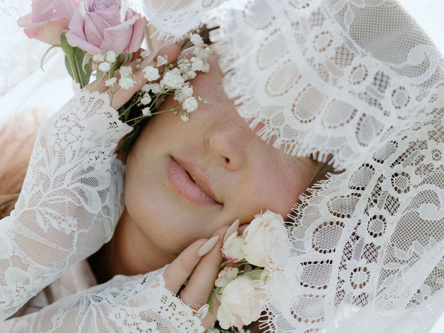 Woman in white lace hat adorned with pink roses and baby's breath, resting peacefully with hands near face.