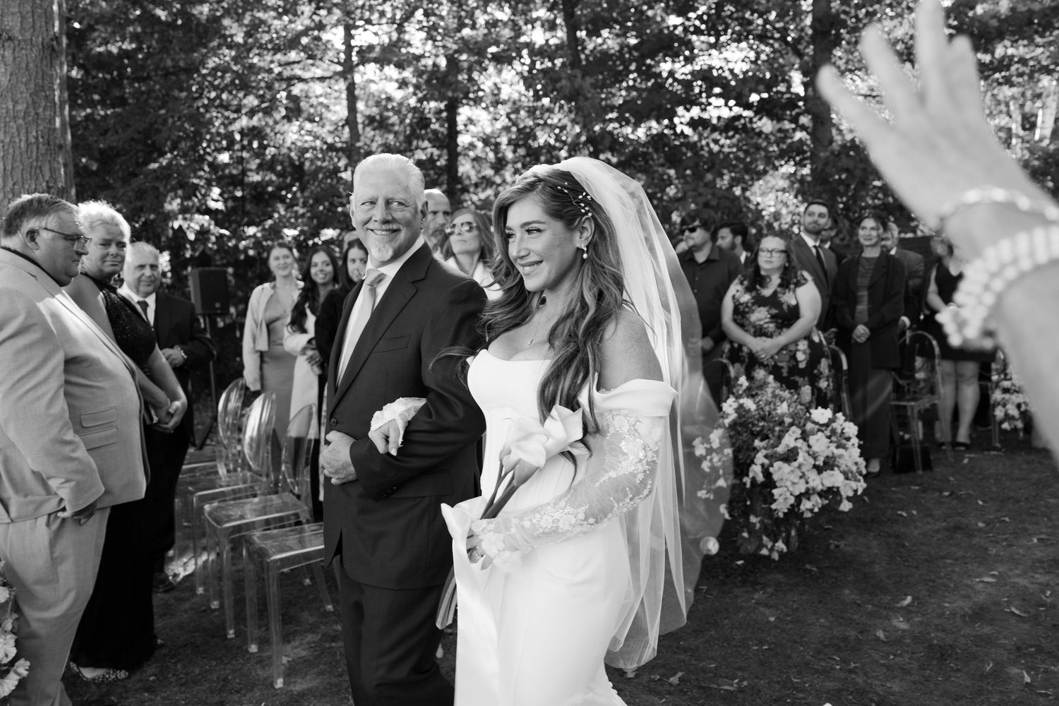 Bride in white gown and veil walks down outdoor aisle with older man in dark suit at wedding ceremony.