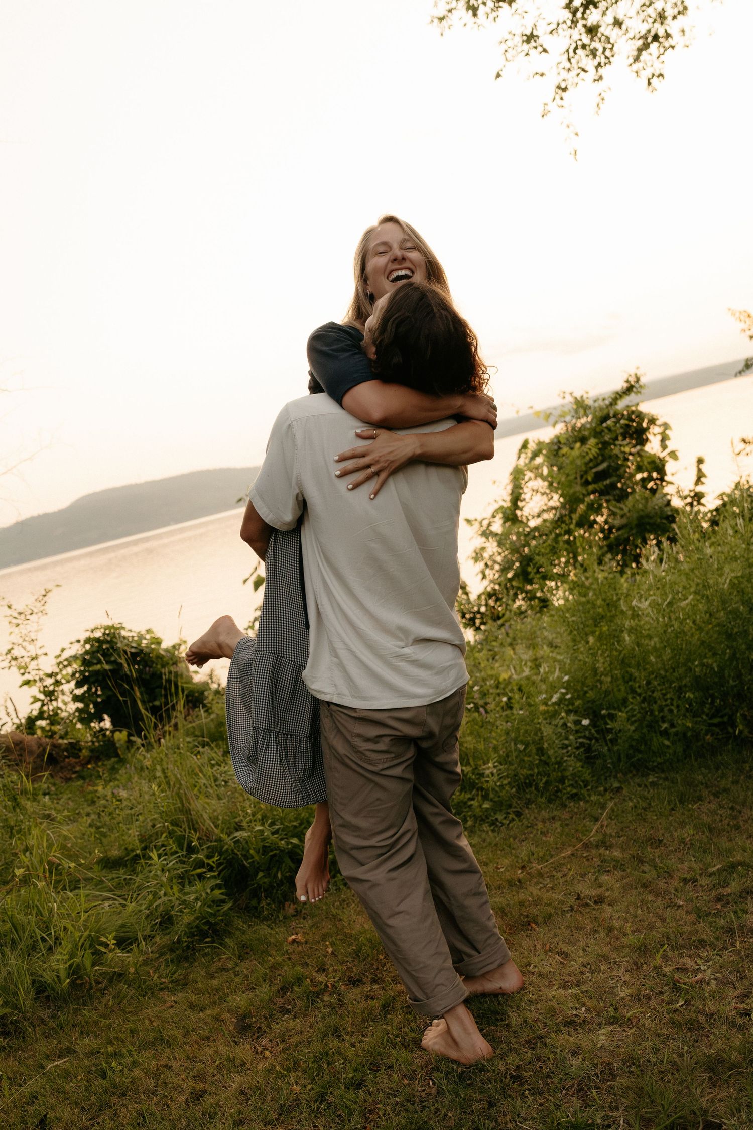 A joyful couple embraces outdoors near a lake, with the woman laughing as the man lifts her up.