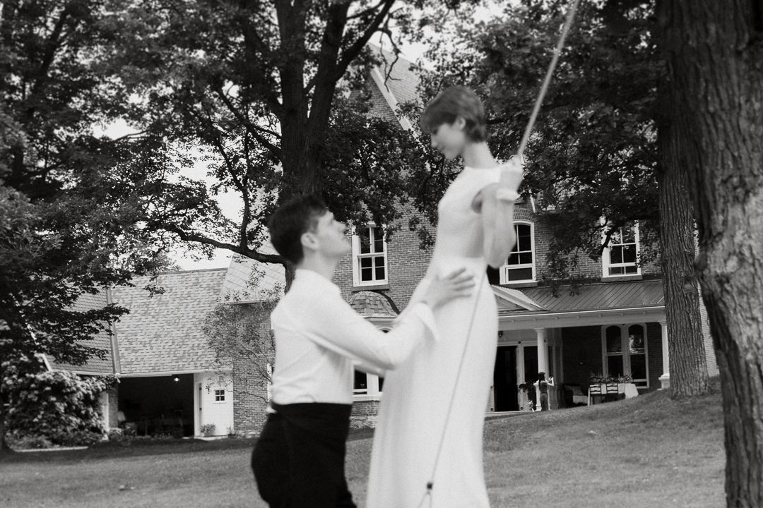Black and white photo of a groom lifting his bride in a white dress outdoors near trees and a house.