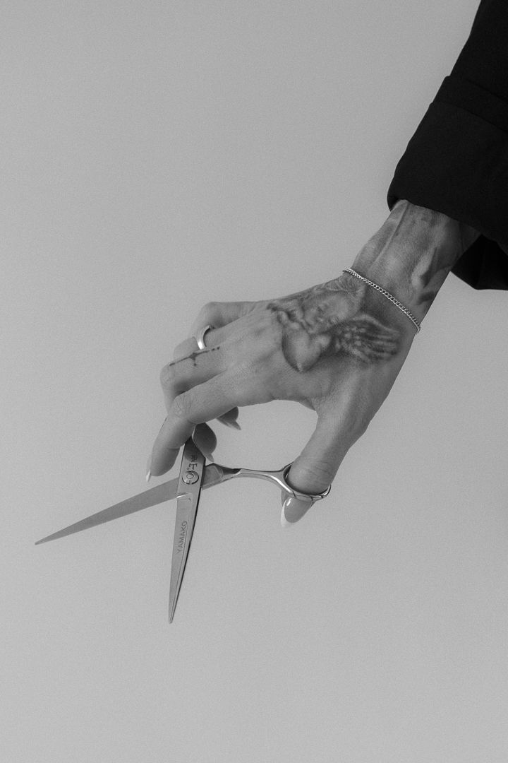 Close-up black and white photo of a tattooed hand with rings and bracelet holding sharp scissors.