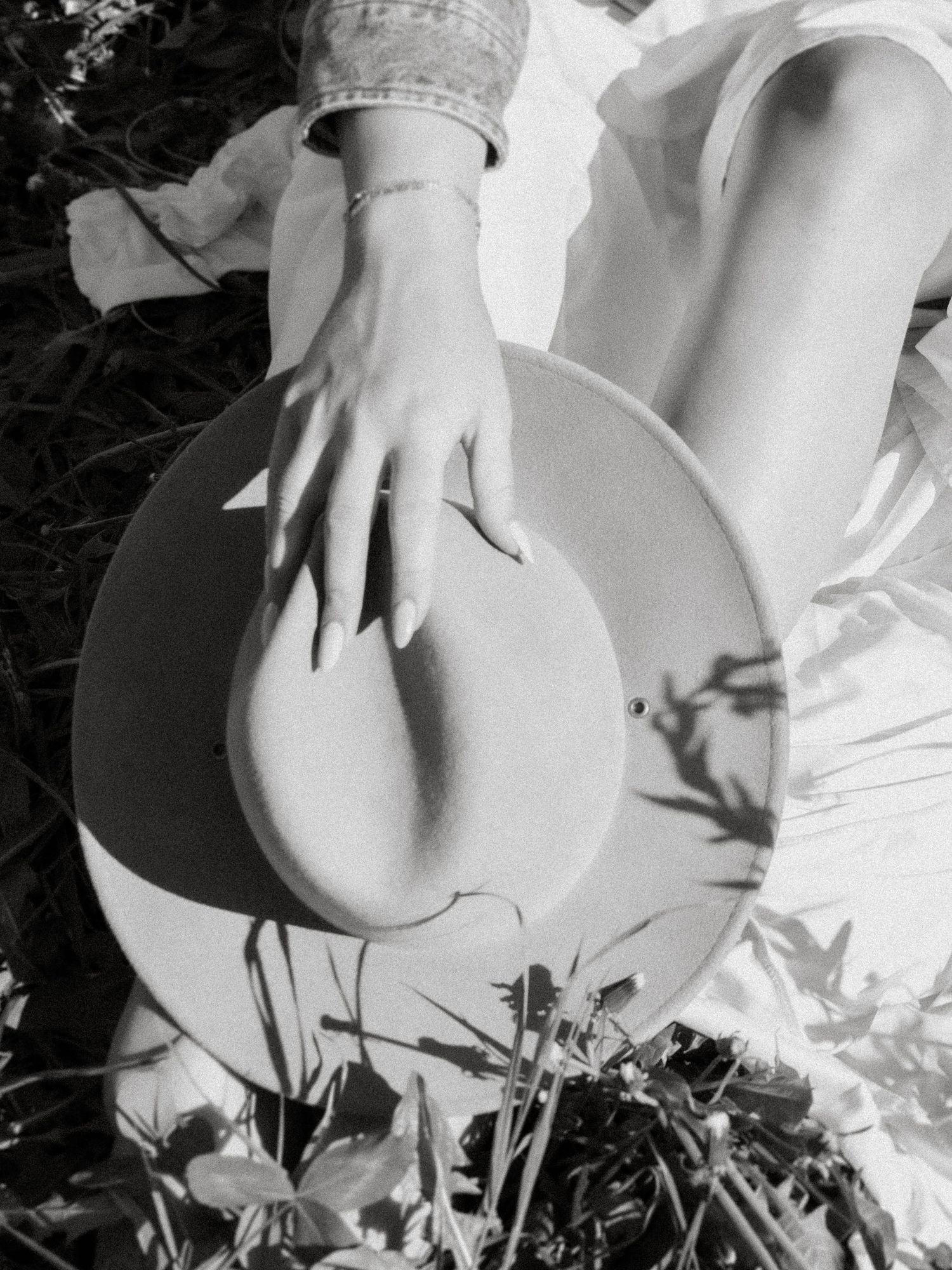 Black and white photo of a woman in white dress holding a wide-brim hat while sitting in grass.