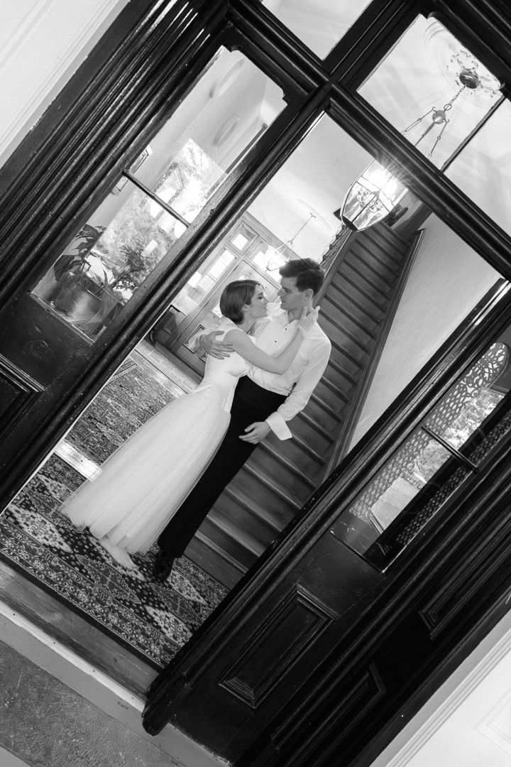 Black and white wedding photo of bride and groom embracing near elegant staircase, viewed through glass doors.