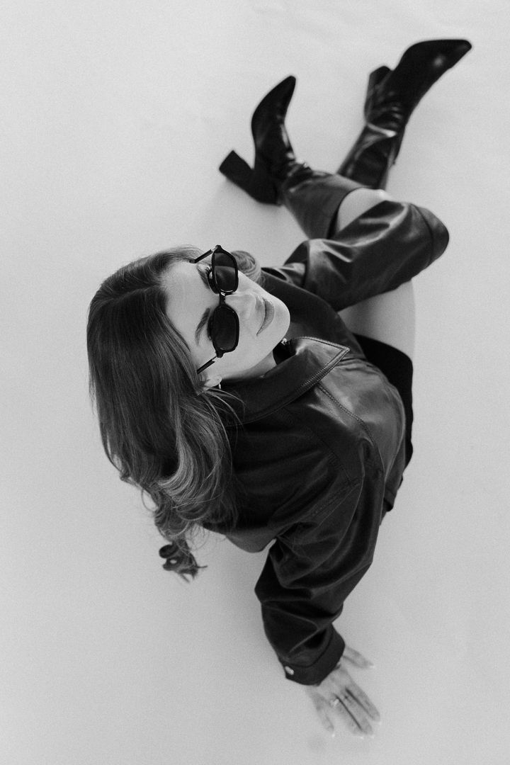 Black and white photo of a woman in leather jacket, pants, and boots sitting on floor wearing sunglasses.