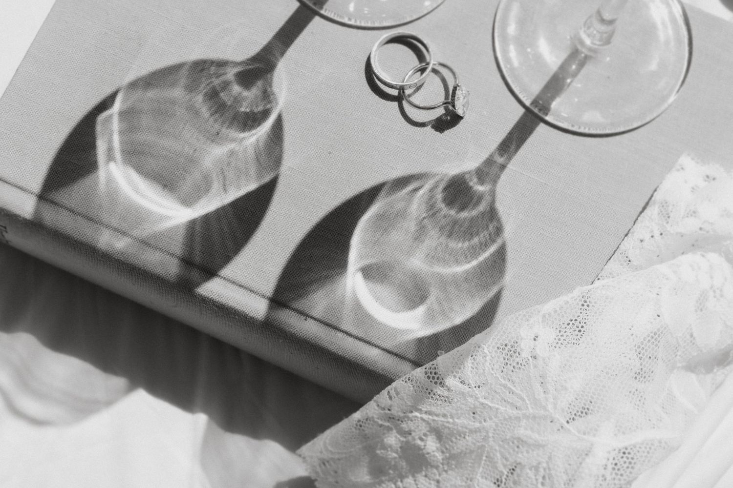 Black and white photo of two wine glasses casting shadows on a surface with wedding rings nearby.