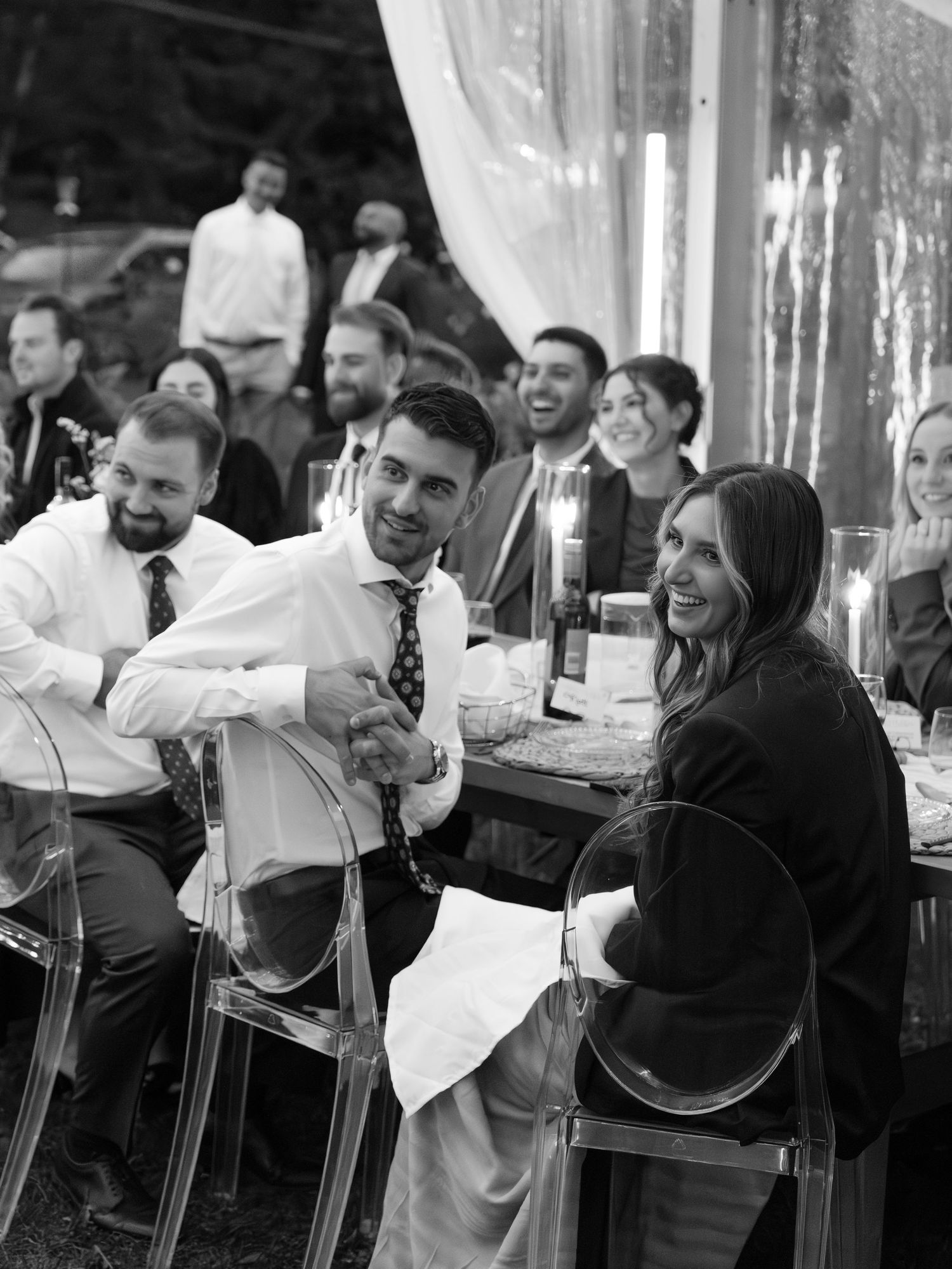 Black and white wedding reception photo of guests laughing and smiling at a candlelit dinner table.