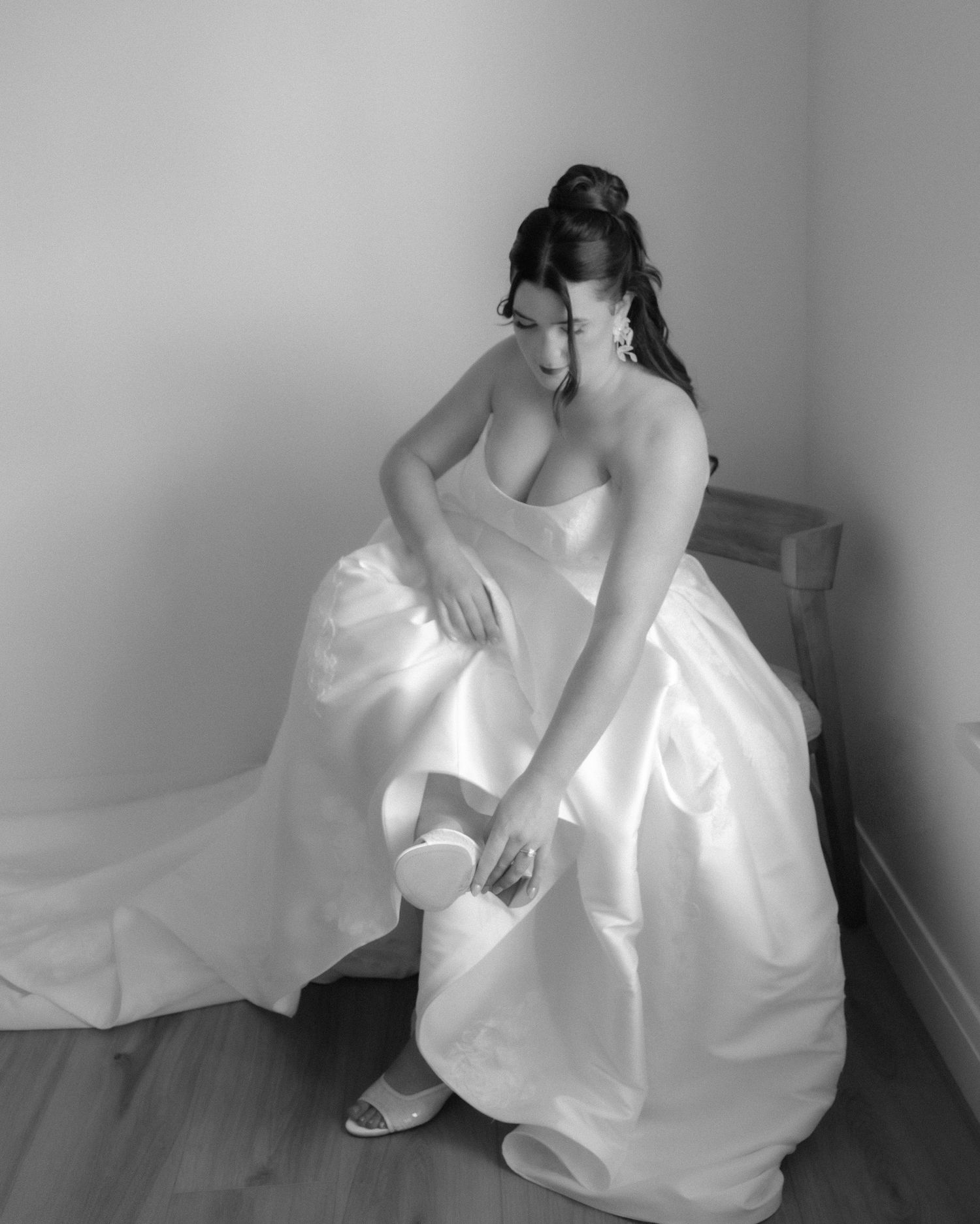 Black and white photo of a bride in a strapless ball gown adjusting her shoe while seated.
