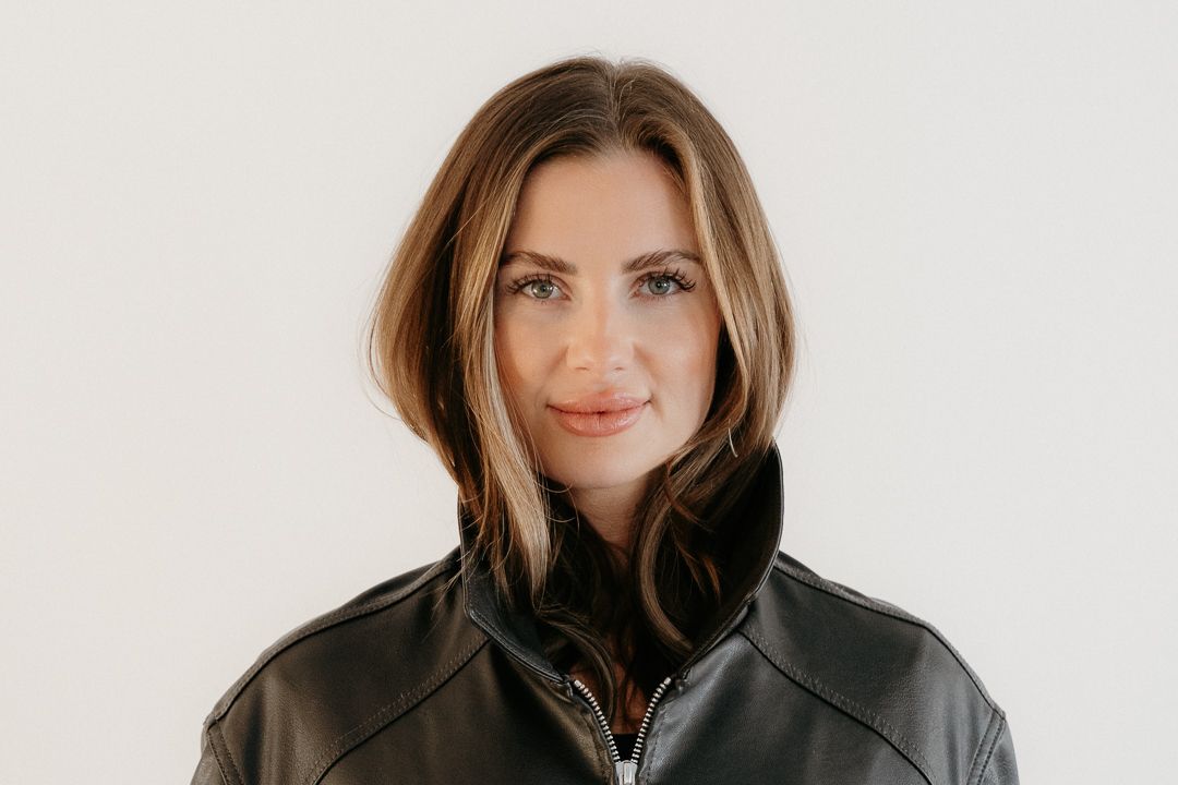 Woman in black leather jacket with highlighted brown hair smiling confidently against a white background.