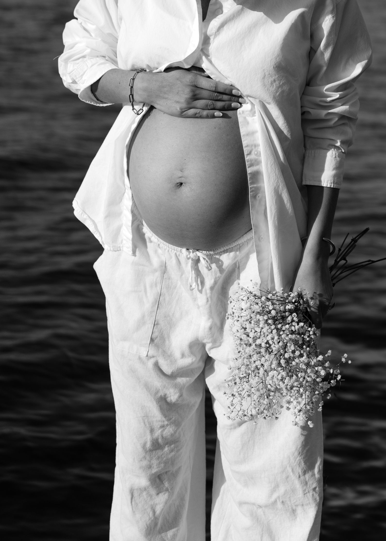 Black and white photo of pregnant woman in white linen outfit holding wildflower bouquet by waterside.