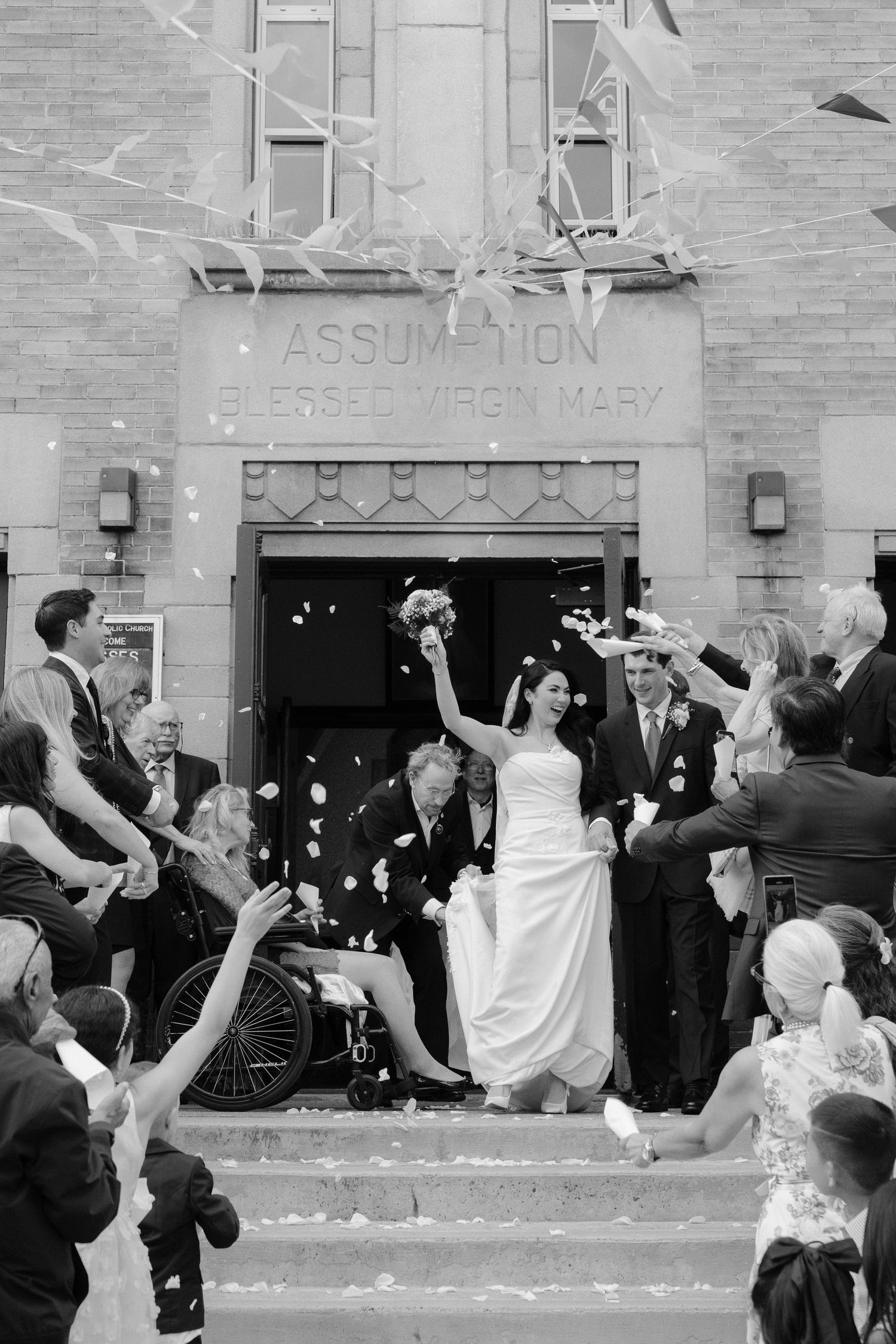 Black and white photo of a bride and groom exiting Assumption Blessed Virgin Mary church amid a shower of confetti.