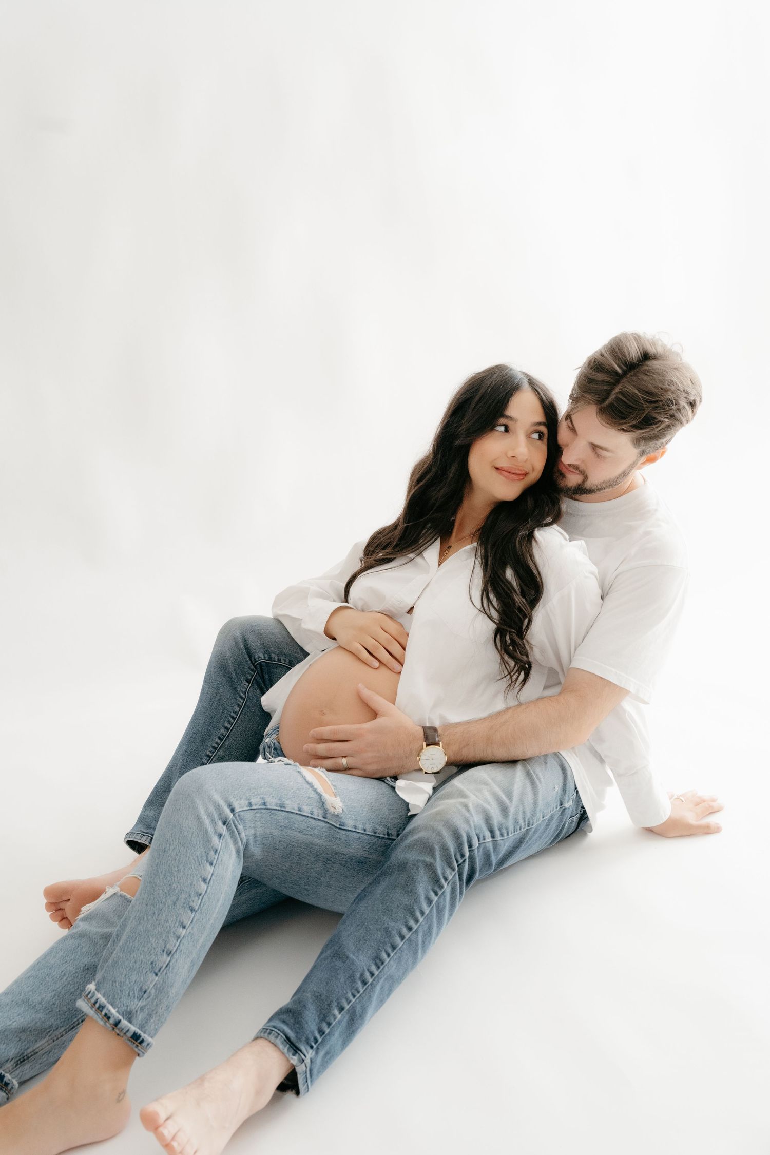 A couple sits on a white studio floor, man embracing pregnant woman in white shirt and jeans, both smiling warmly.