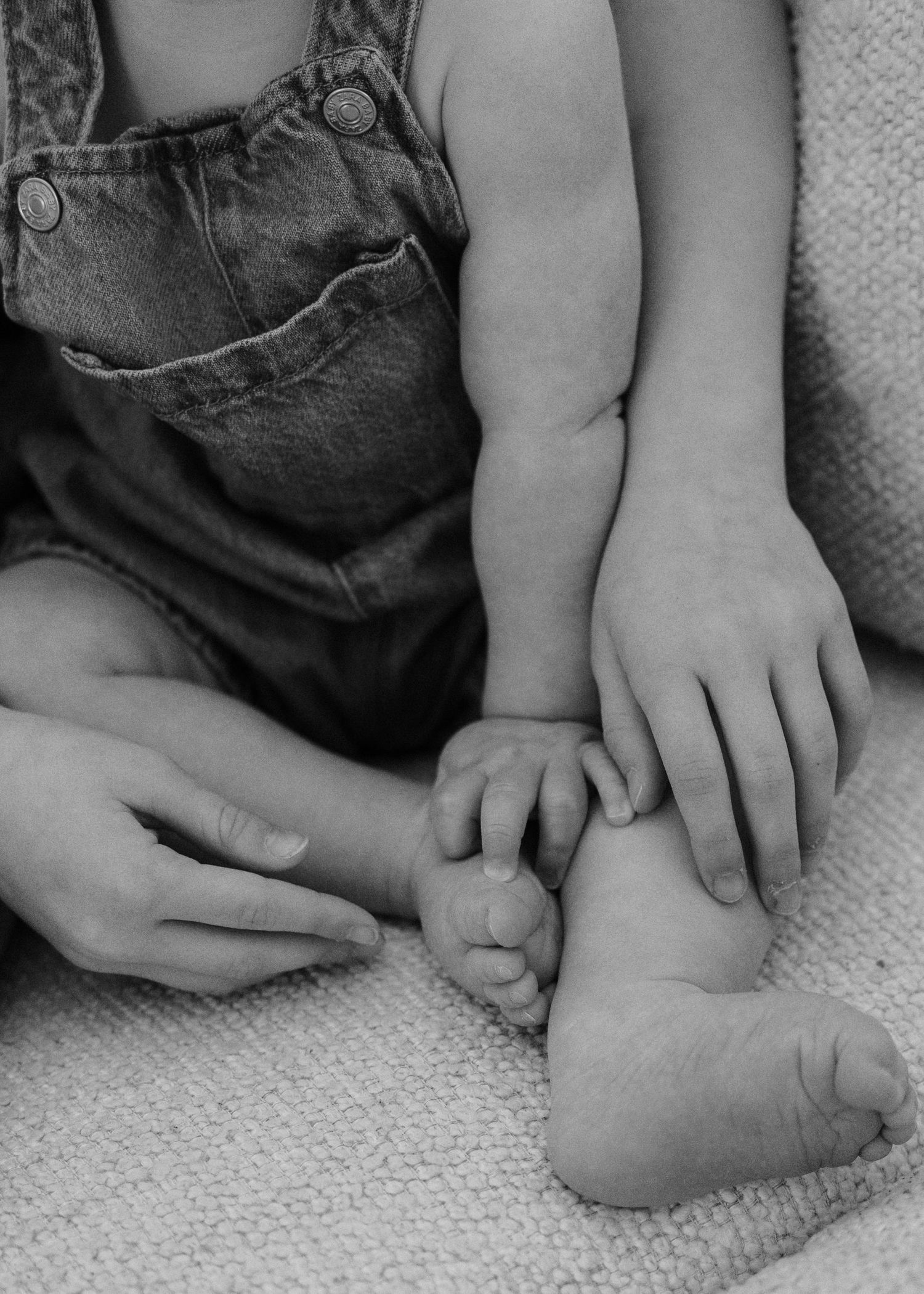 Black and white photo of a baby's tiny foot held gently by an adult hand, with a toddler in denim overalls nearby.