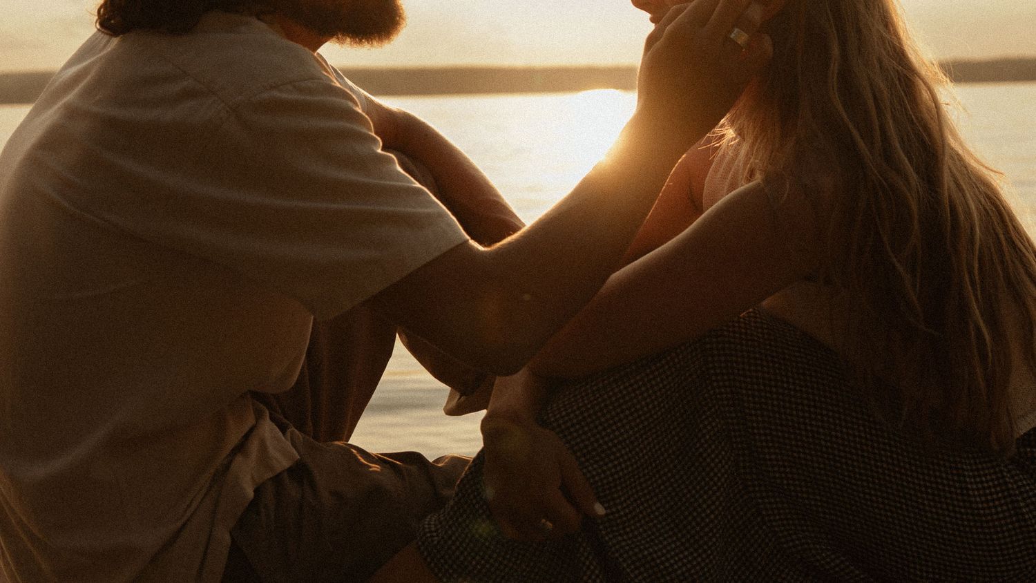 Couple sitting together at sunset by the water, sharing an intimate moment in warm golden light.