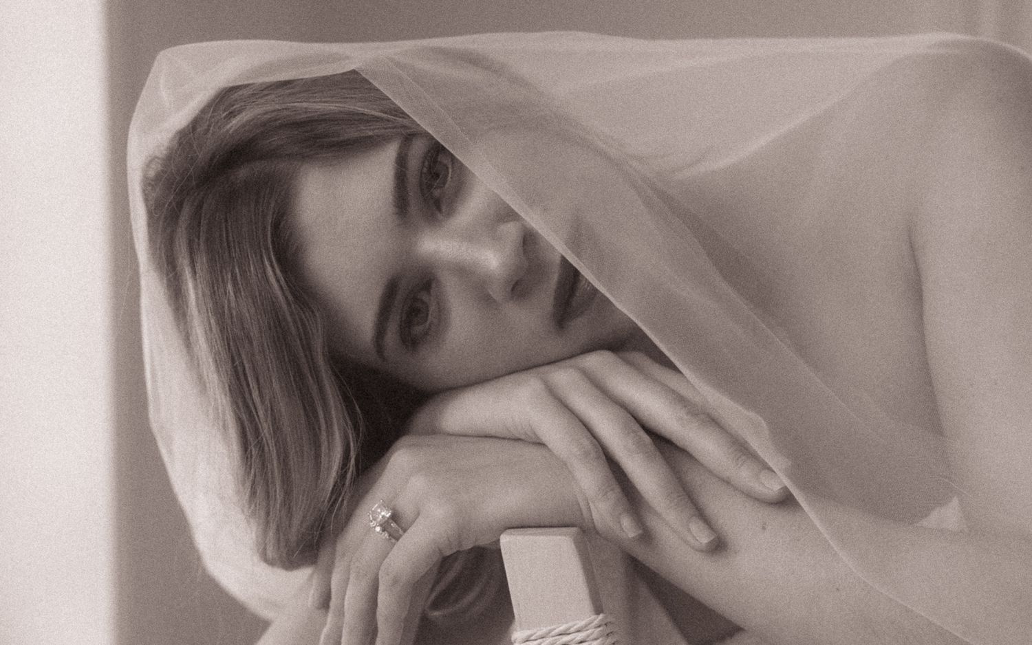 Sepia-toned close-up of bride with veil draped over head, hands folded near face showing delicate ring.