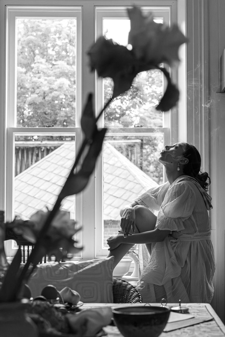 Black and white photo of a pregnant woman in a robe sitting by a sunlit window, surrounded by flowers and fruit.