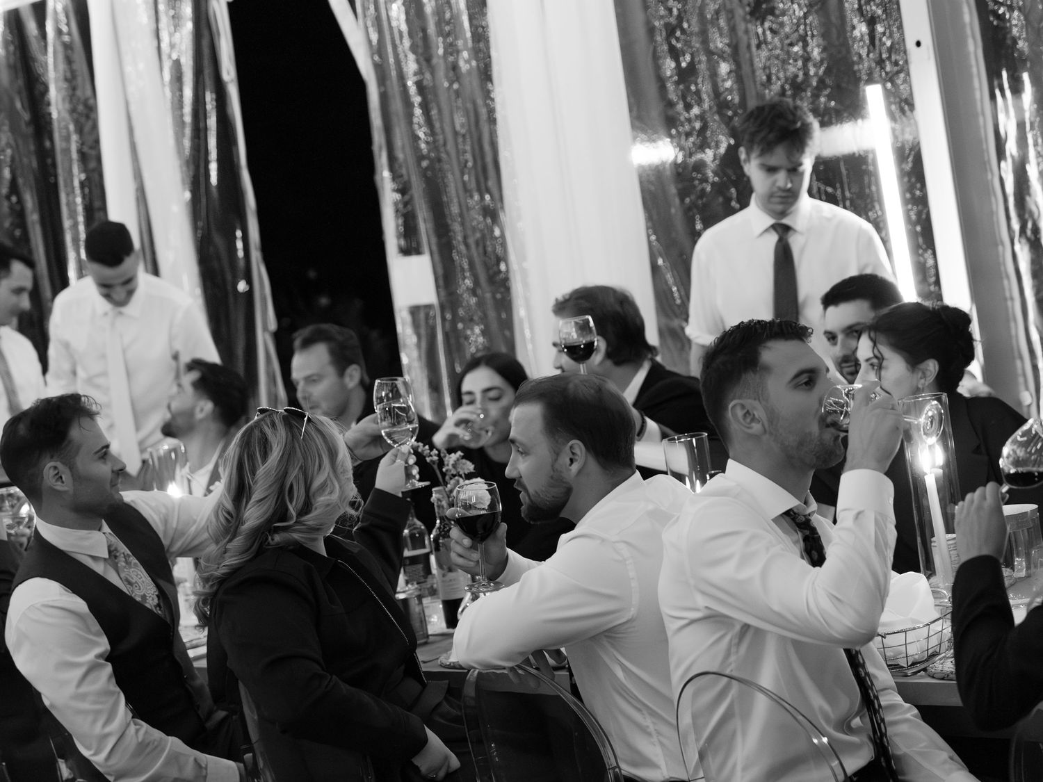 Black and white photo of wedding reception guests seated at tables under shimmering backdrop.
