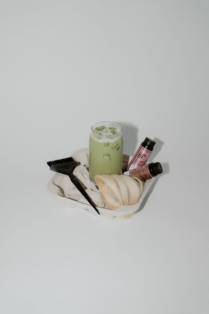 Hair dye tools with gloves, brush, color bottle, and iced matcha latte arranged on white fabric.