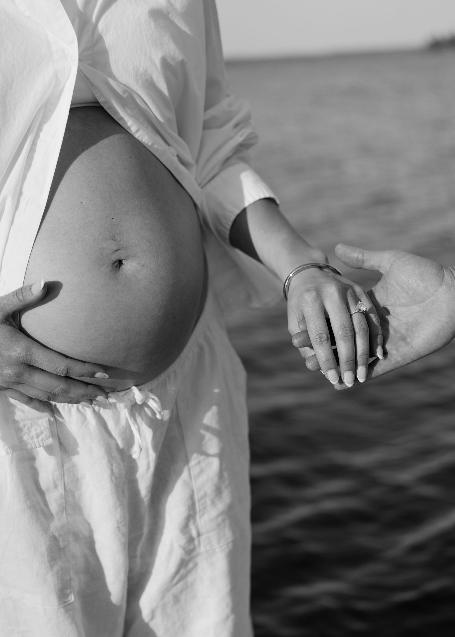Black and white maternity photo of pregnant couple holding hands near sparkling water backdrop.